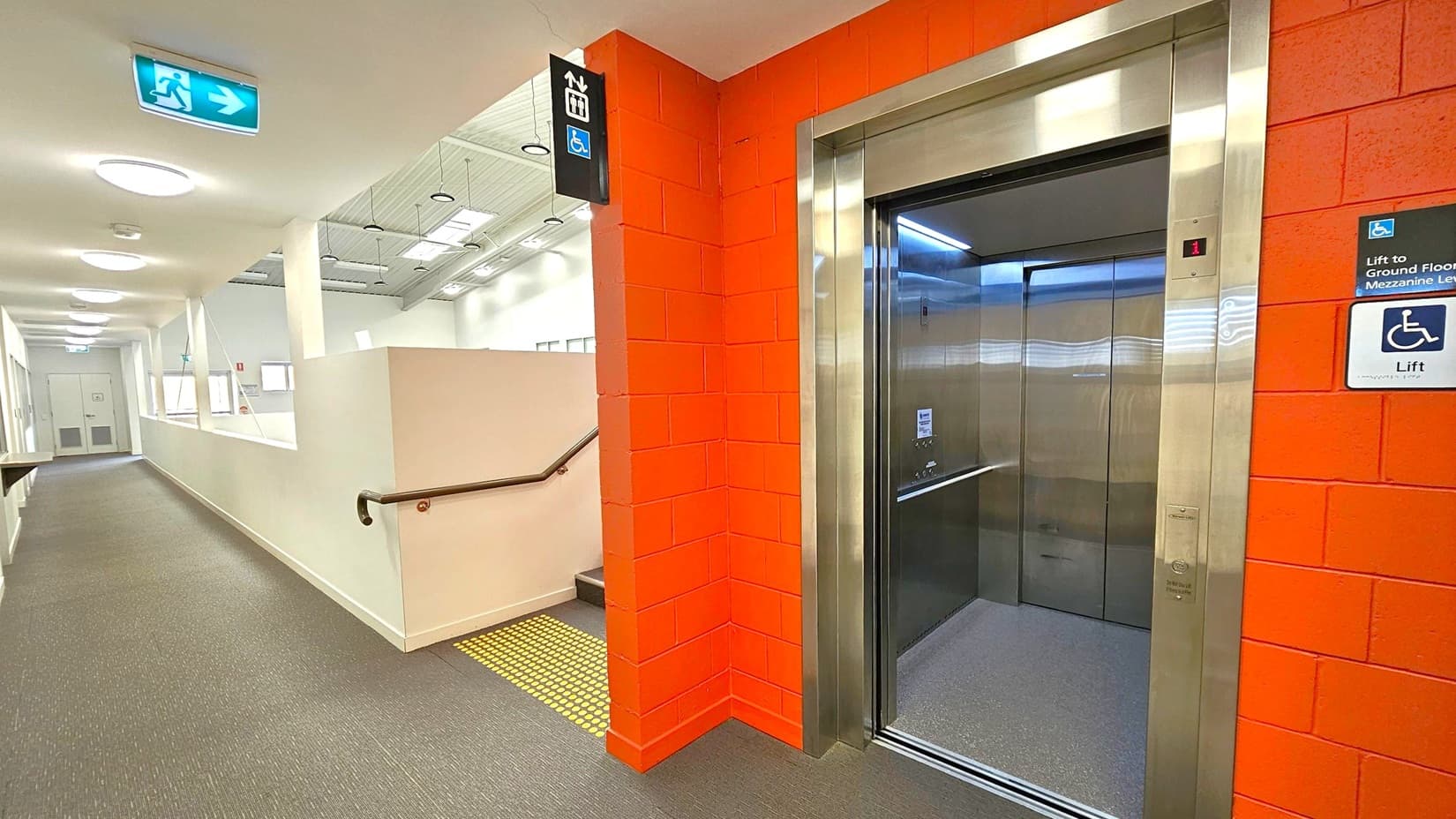 A corridor with a grey carpeted floor and white walls. On the right, there's an open lift with a stainless steel frame set into a red brick wall. Above the lift, a sign indicates accessibility. To the left, a series of round ceiling lights illuminate the hallway, which leads to a door at the end. An emergency exit sign with a running figure and arrow is mounted on the ceiling. A handrail runs along the wall, and a small staircase is visible near the lift.