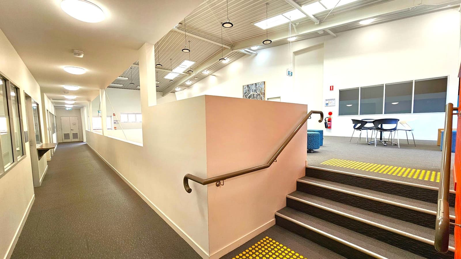A corridor with a brown carpeted floor on the left, featuring a row of ceiling lights and windows along the wall. On the right, a short flight of stairs with a handrail leads up to a carpeted area with a table and chairs. The ceiling is high with exposed beams and hanging lights. Skylights allow natural light in. A fire extinguisher is mounted on the wall near the stairs, and a piece of artwork is displayed above a blue cushioned seat.