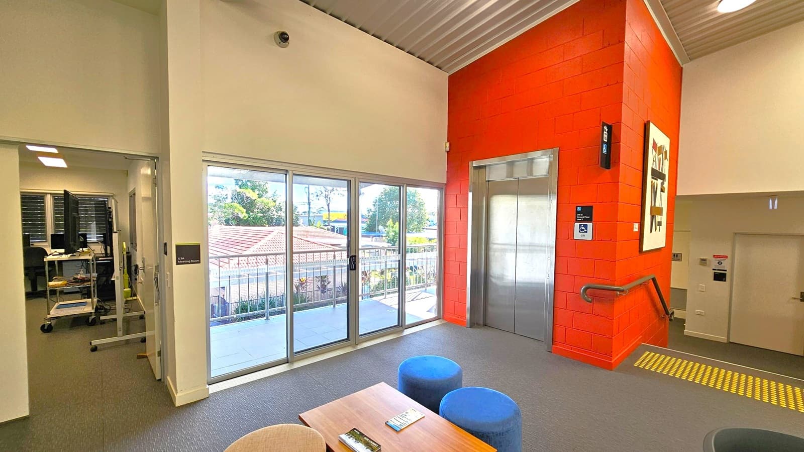 A room with a grey carpeted floor and a corrugated metal ceiling. On the right, there's a red brick wall with a lift and a handrail leading to stairs. A piece of abstract art is mounted on the wall. In the centre, a set of large glass sliding doors opens to a balcony with a view of trees and rooftops. On the left, an open doorway leads to a room with computer equipment. In the foreground, a wooden table with brochures and two blue stools are visible.