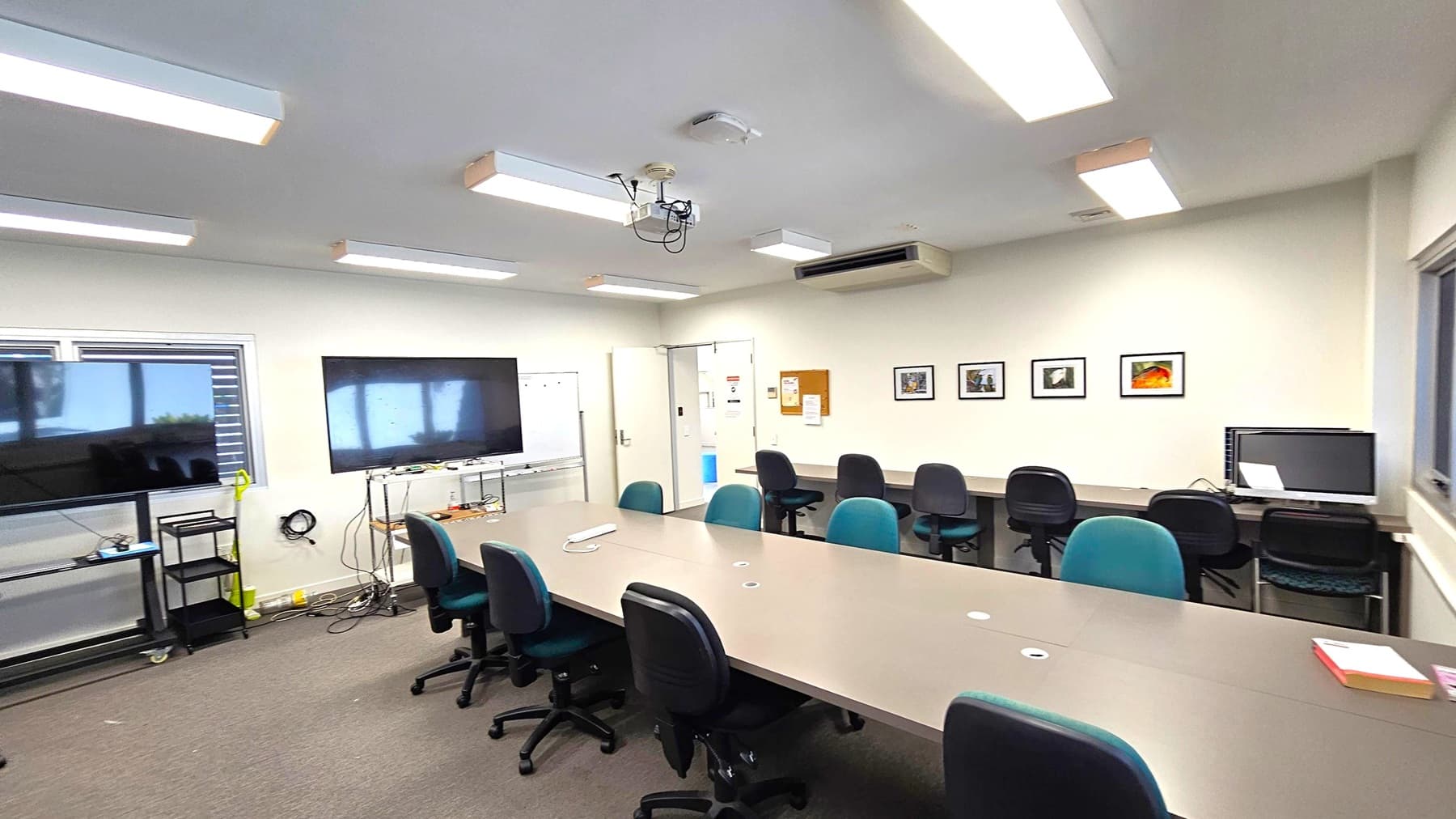 A conference room with a long table surrounded by office chairs. On the left, there are two large screens, one on a stand and another mounted on the wall, with a whiteboard nearby. The right wall features framed pictures and a computer on a desk. The ceiling has several fluorescent lights and a projector. The floor is carpeted, and there are windows with blinds on the left side.
