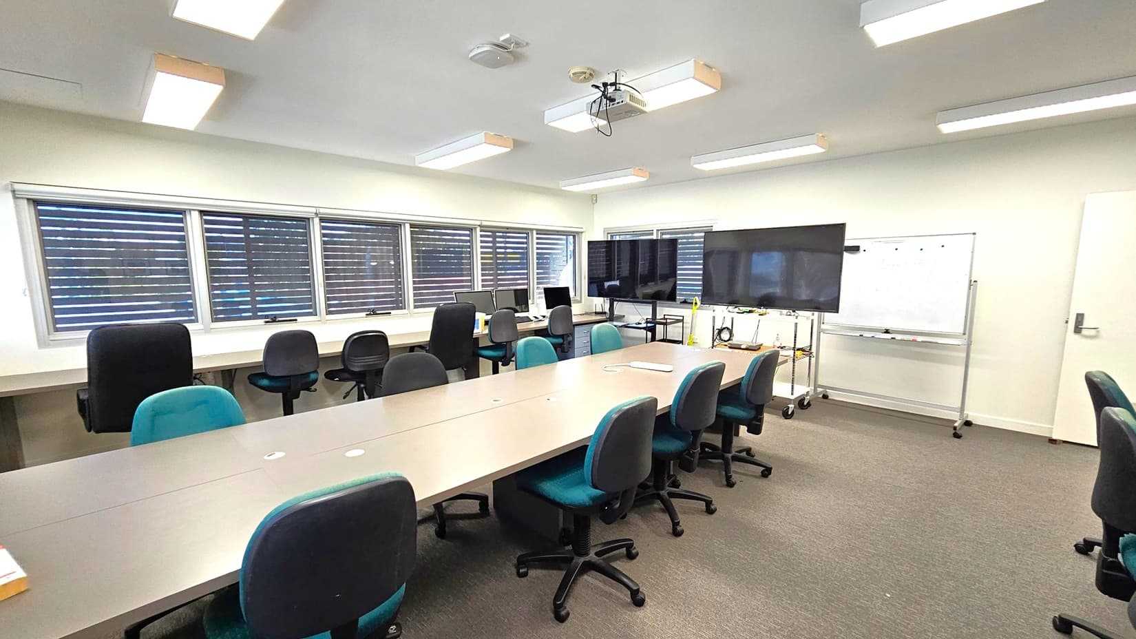 A conference room with a long table in the centre surrounded by office chairs. On the left, there are windows with blinds and a row of desks with computers and chairs. On the right, a large monitor is mounted on a stand, and a whiteboard is nearby. The ceiling has several fluorescent lights and a projector. The floor is carpeted in a brown and green pattern.