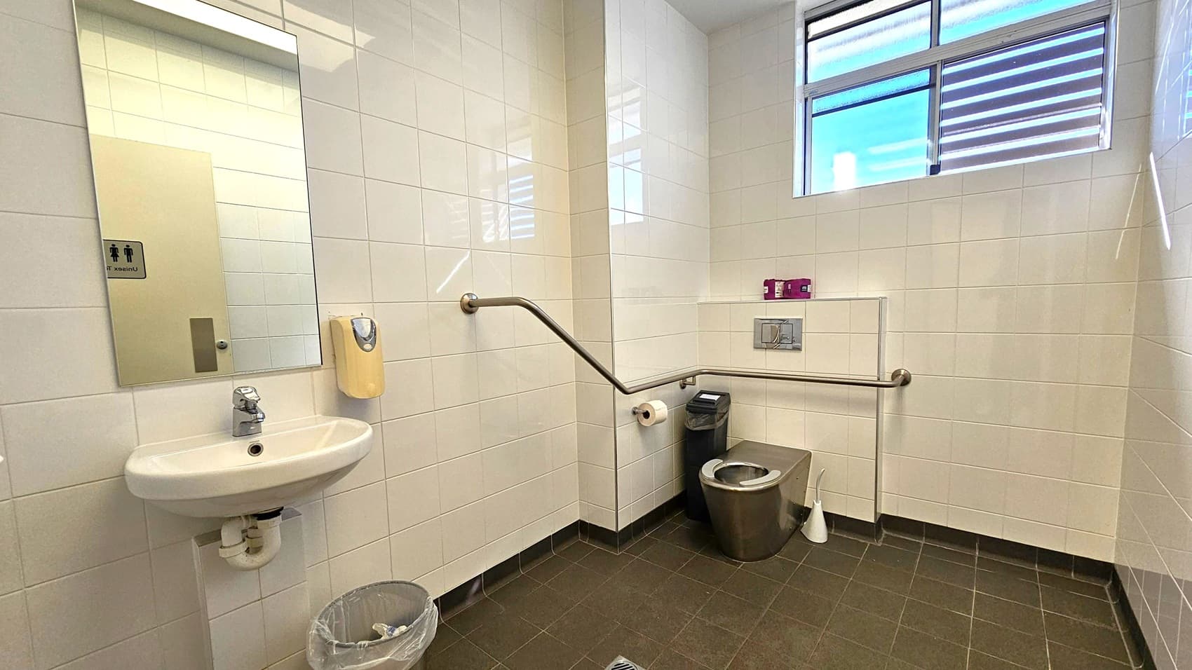 A public restroom with white tiled walls and a dark tiled floor. On the left, there's a sink with a mirror above it and a soap dispenser mounted on the wall. Below the sink is a small waste bin. On the right, a stainless steel toilet with a toilet roll holder and a sanitary bin nearby. A grab bar runs along the wall for accessibility. Above, a window with horizontal blinds allows natural light into the room.