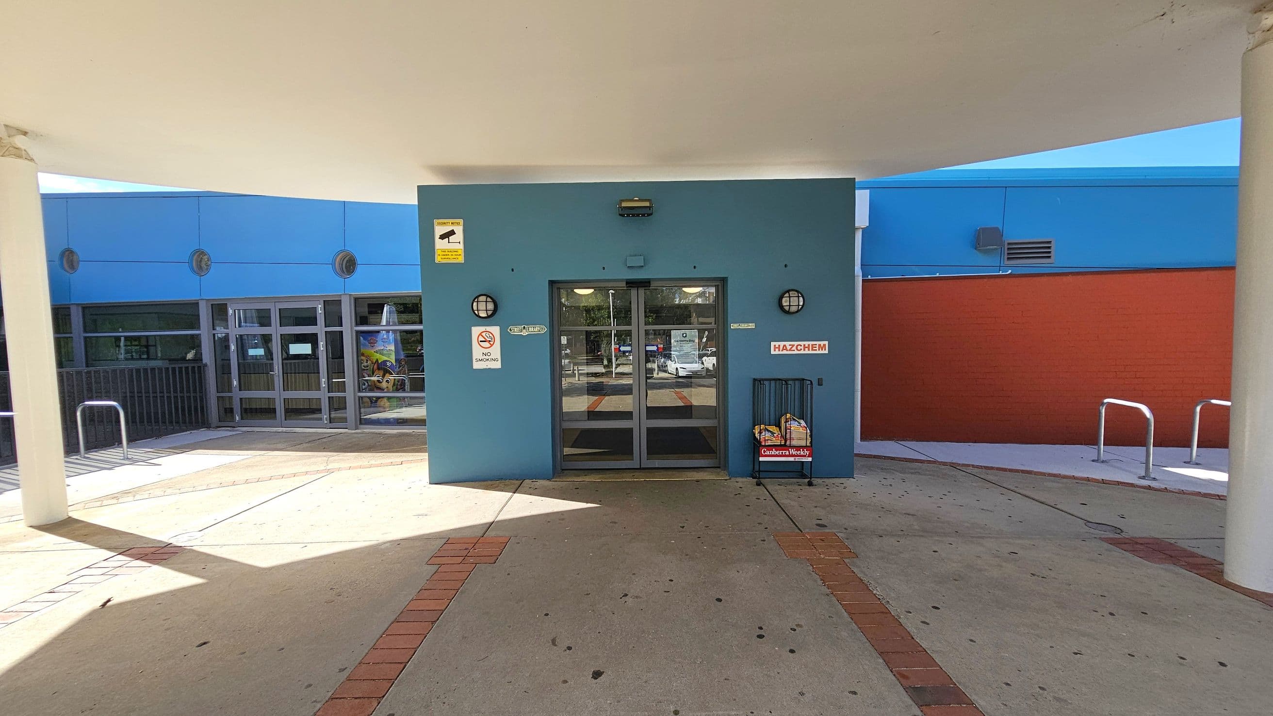 A building entrance with a blue facade and glass double doors in the centre. Above the doors, there's a security camera sign. To the left, a glass window and a metal railing are visible. On the right, a red brick wall and metal bike racks are present. A newspaper stand with "Canberra Weekly" is near the entrance. The floor is concrete with brick accents, and the ceiling is white with a curved design.