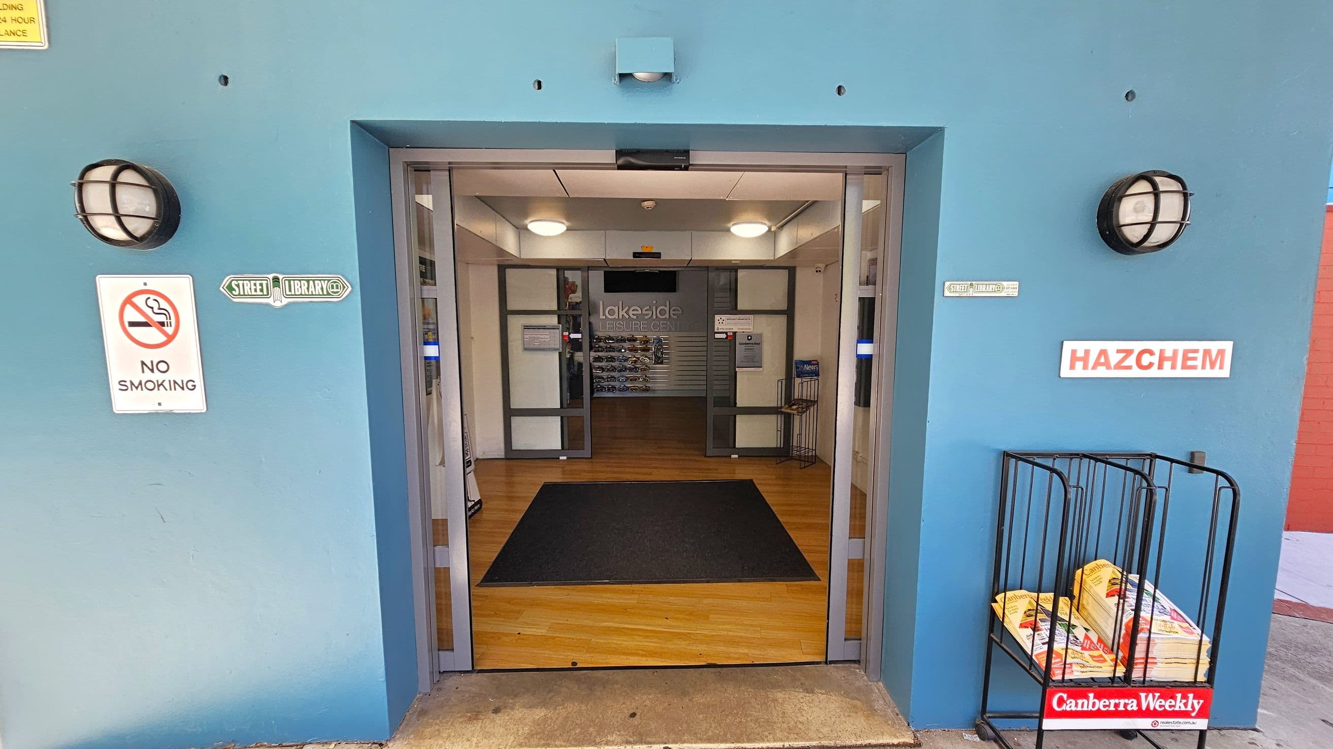 A blue entrance with double glass doors leading into a building with a wooden floor. On the left, a "No Smoking" sign and a "Street Library" sign are mounted on the wall. On the right, a "HAZCHEM" sign is visible above a rack holding copies of "Canberra Weekly". Two round wall lights are positioned on either side of the entrance. Inside, the word "Lakeside" is visible on the back wall.