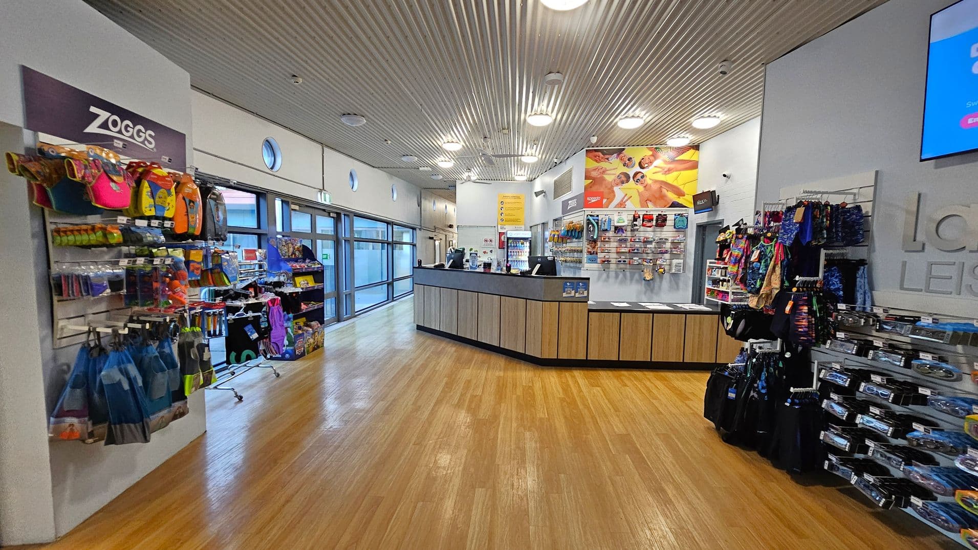 A leisure centre shop with a wooden floor and a corrugated metal ceiling. On the left, shelves display swimming gear and accessories with a "Zoggs" sign above. The centre features a reception counter with promotional materials and a display of goggles and swimwear behind it. On the right, more shelves hold colourful swimwear and accessories. Large windows line the left wall, allowing natural light to enter.
