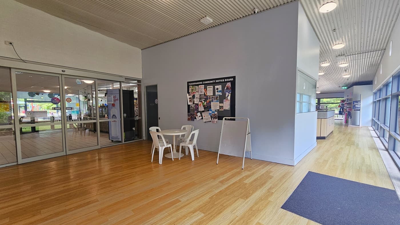 A spacious indoor area with wooden flooring and a corrugated ceiling. On the left, there are glass sliding doors leading to another room with visible decorations. In the centre, a small round table with three white plastic chairs is placed against a wall featuring a community notice board filled with various flyers and notices. On the right, a counter and a corridor with large windows allowing natural light to enter. Ceiling lights are evenly spaced along the corridor.