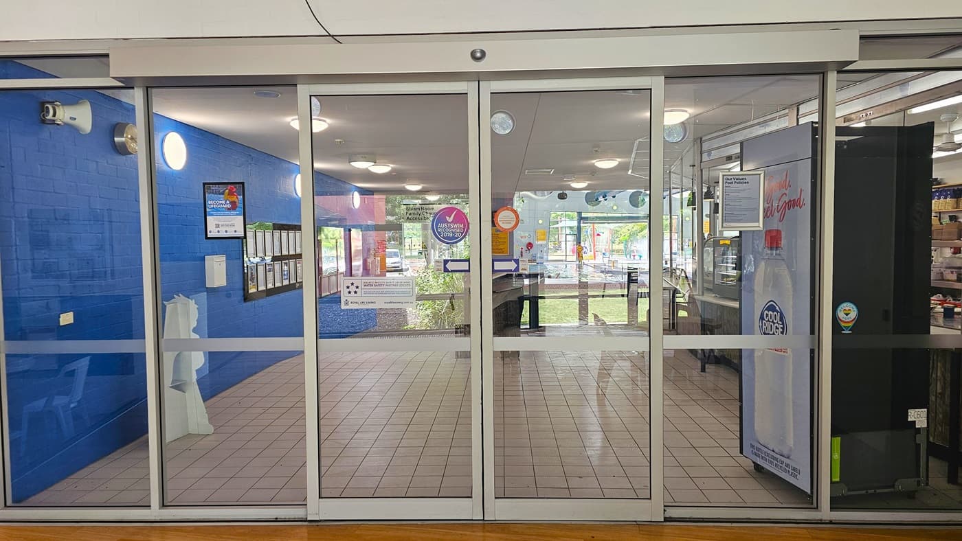 Glass sliding doors with a metal frame, leading into a building. The left side has a blue wall with framed notices and a white dispenser. The right side shows part of a vending machine with a "Cool Ridge" label. Inside, the floor is tiled, and there are several circular ceiling lights. Through the glass, tables and chairs are visible, along with more notices and a view of greenery outside. Stickers and signs are on the doors.