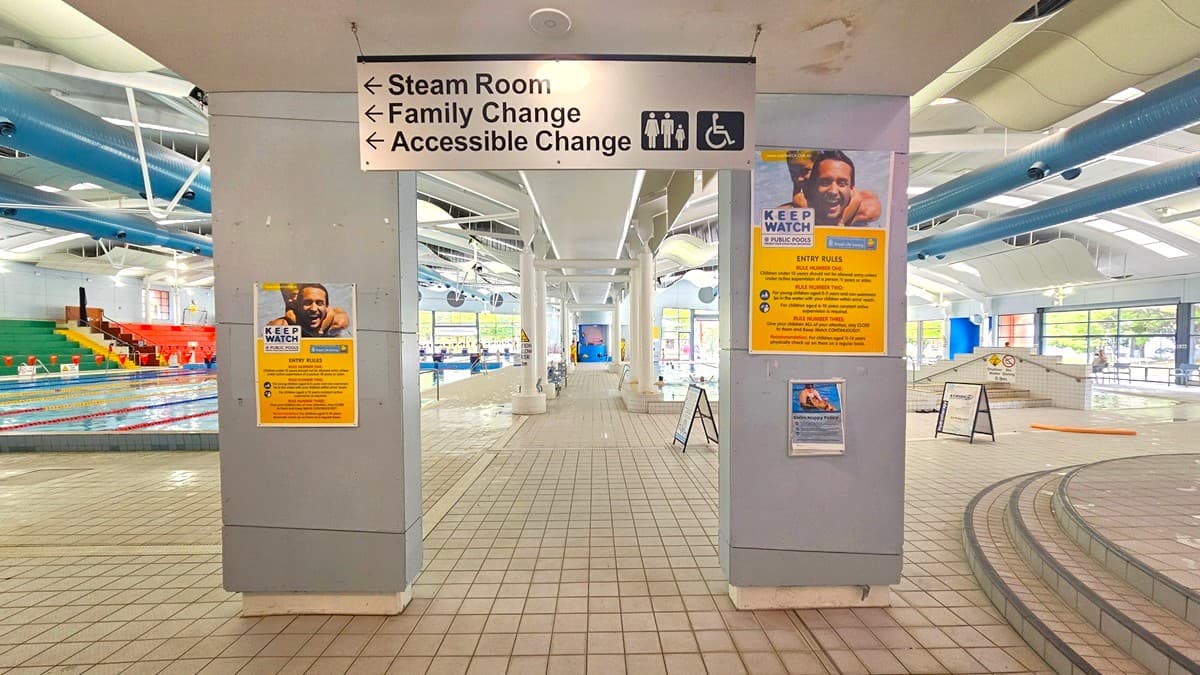 A swimming pool area with tiled flooring and a high ceiling featuring exposed pipes. In the centre, a sign indicates directions to the steam room, family change, and accessible change. On the left, a pool with colourful seating in red, yellow, and green. On the right, a ramp and steps lead to another pool area. Posters with safety information are displayed on the columns. Large windows in the background allow natural light to enter.