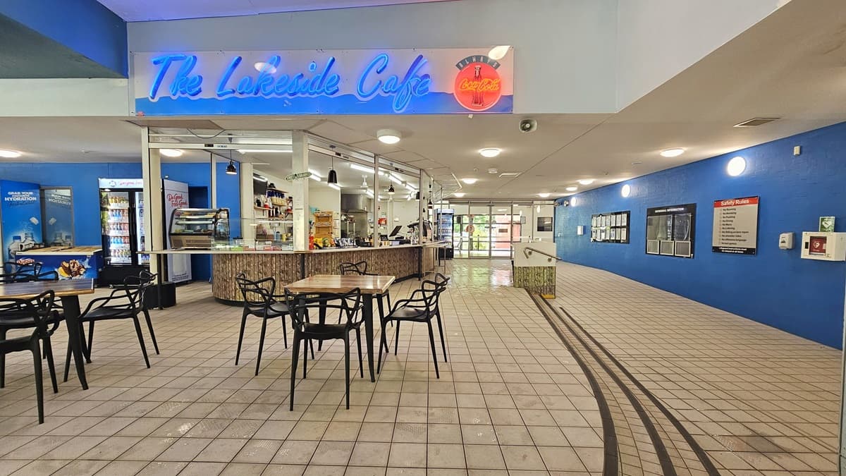 A café with a neon sign reading "The Lakeside Cafe" is on the left. It features a counter with various items and a display case. Several tables and chairs are in front of the counter. The floor is tiled, and the ceiling has recessed lighting. On the right, a blue wall displays information boards and signs. A ramp with a railing leads towards a glass entrance in the background.