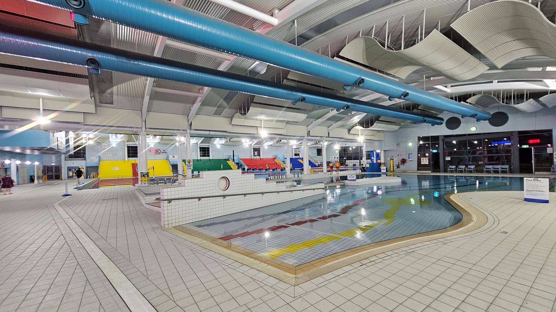 An indoor swimming pool area with a tiled floor and a pool featuring colourful lane markings. On the left, there are steps leading to a yellow wall, with seating in green, red, and blue. The ceiling has large blue pipes and wavy acoustic panels. On the right, large windows and doors are visible, with tables and chairs nearby. The lighting is bright, and the space is spacious and open.