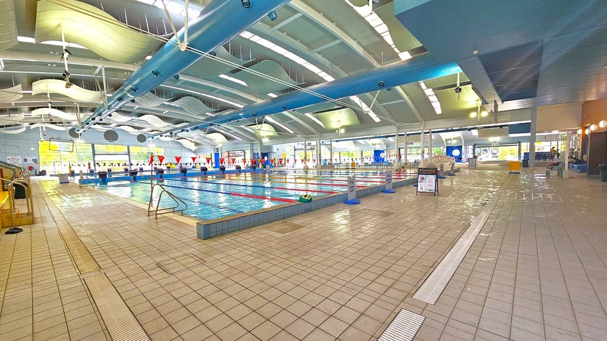 Indoor swimming pool with multiple lanes, marked by red and white flags. The pool is surrounded by a tiled floor. On the left, there are steps leading into the pool. The ceiling features curved, wave-like panels and blue beams. Large windows on the far side allow natural light to enter. The right side has a seating area and informational signs. The space is well-lit with overhead lights.