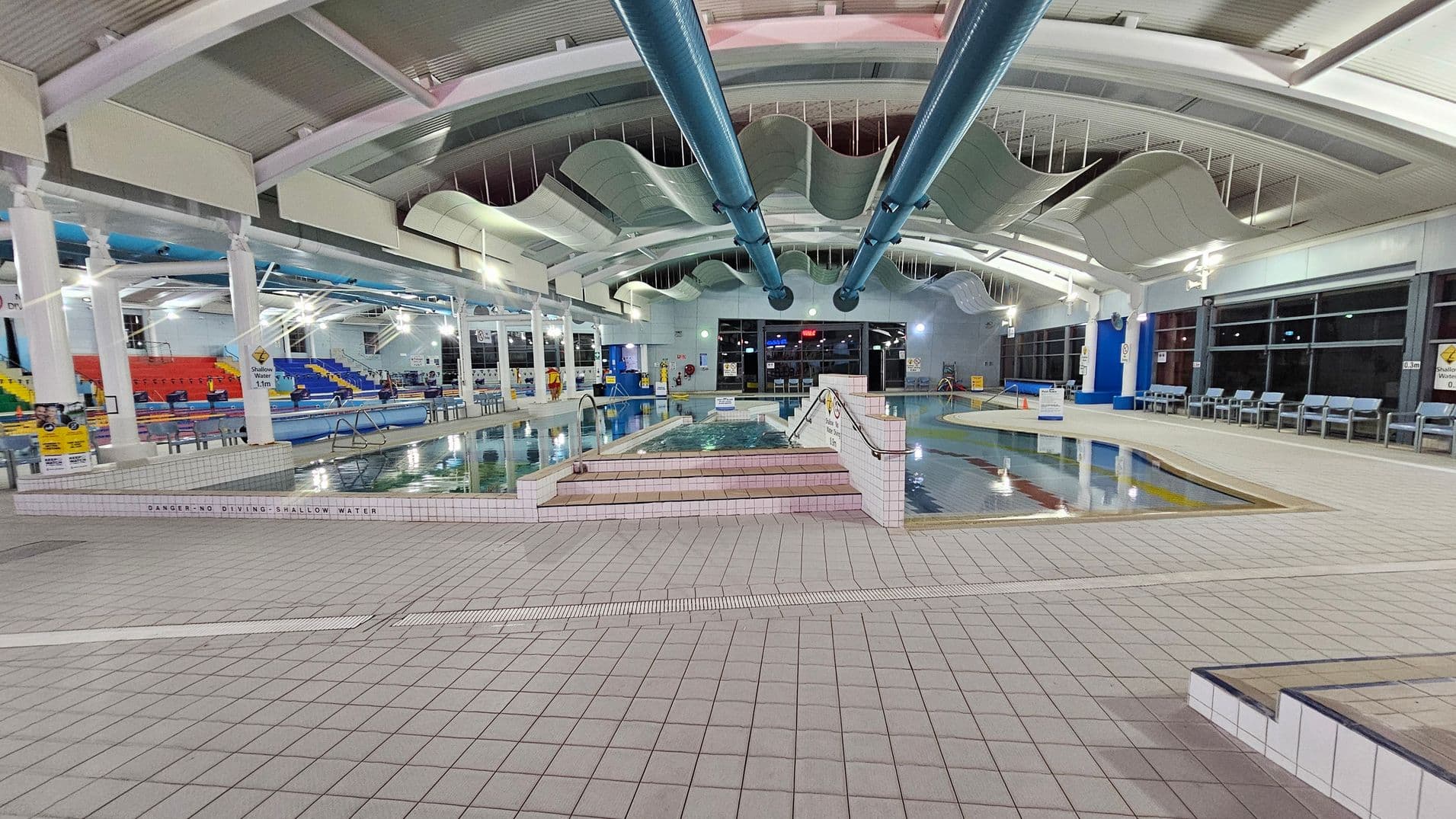 An indoor swimming pool area with a tiled floor and a wavy ceiling design. In the centre, there are two large blue pipes running along the ceiling. The pool is surrounded by a tiled deck, with steps leading into the water. On the left, there are colourful seating areas with red, yellow, and blue seats. On the right, a row of blue chairs lines the wall. Large windows are visible in the background, and various signs are placed around the pool area.