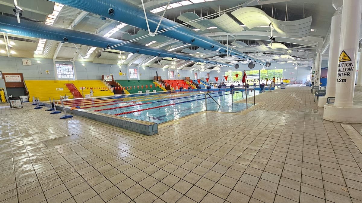 Indoor swimming pool with lanes marked by red and white ropes. On the left, there are yellow and green spectator seats with red stairs. The right side has a tiled pool deck with a caution sign on a pillar. The ceiling features large blue pipes and white curved panels. The floor is covered with light-coloured tiles. Bright overhead lighting and windows provide illumination.