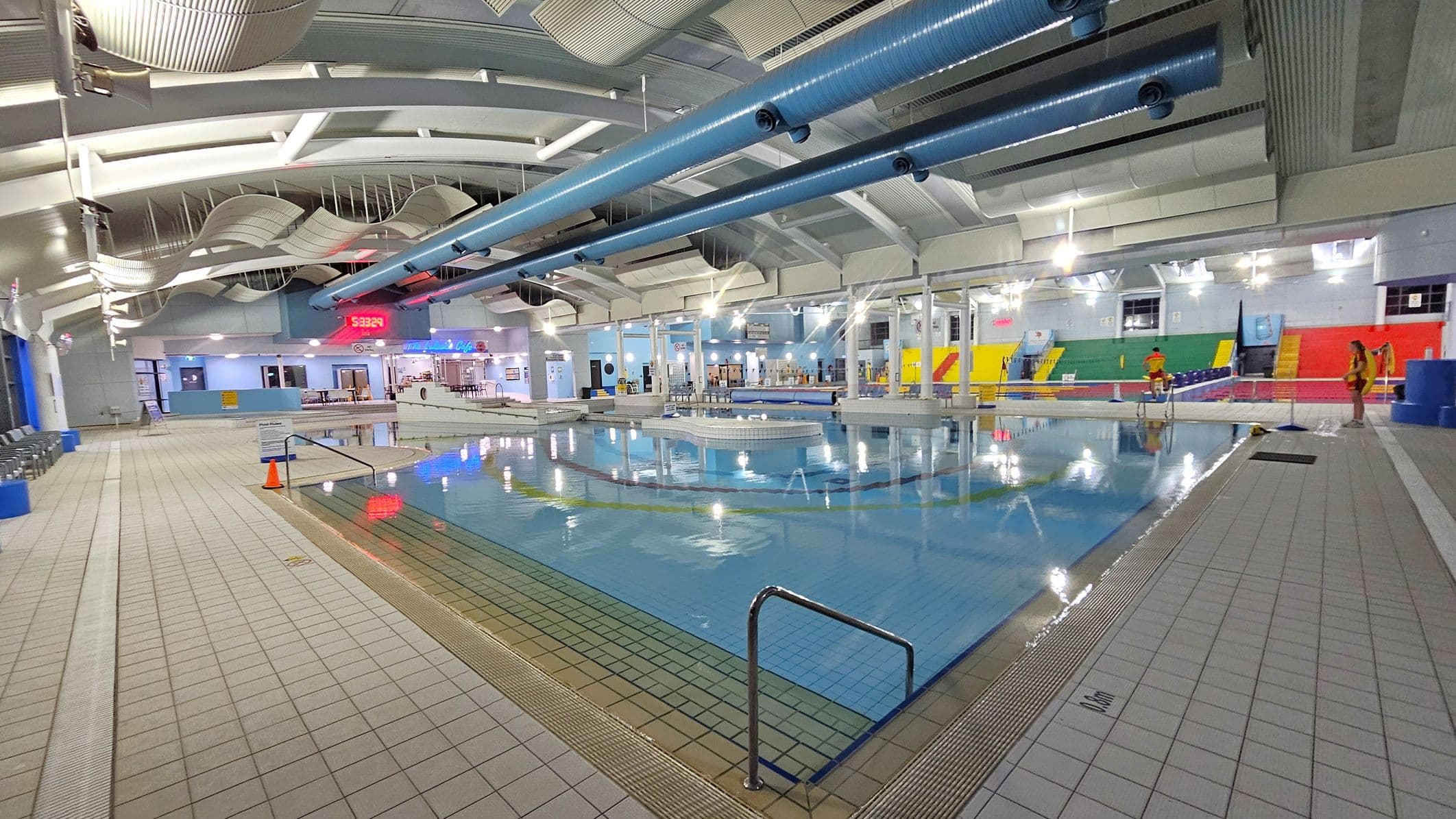 An indoor swimming pool area with a tiled floor surrounding the water. On the left, there are rows of chairs along the wall. The ceiling features curved panels and large blue ventilation ducts. In the centre, a digital clock displays the time. The background includes a café area with tables and chairs. On the right, colourful seating in red, yellow, and green is visible, with a few people standing nearby. The pool has a metal handrail at the edge.