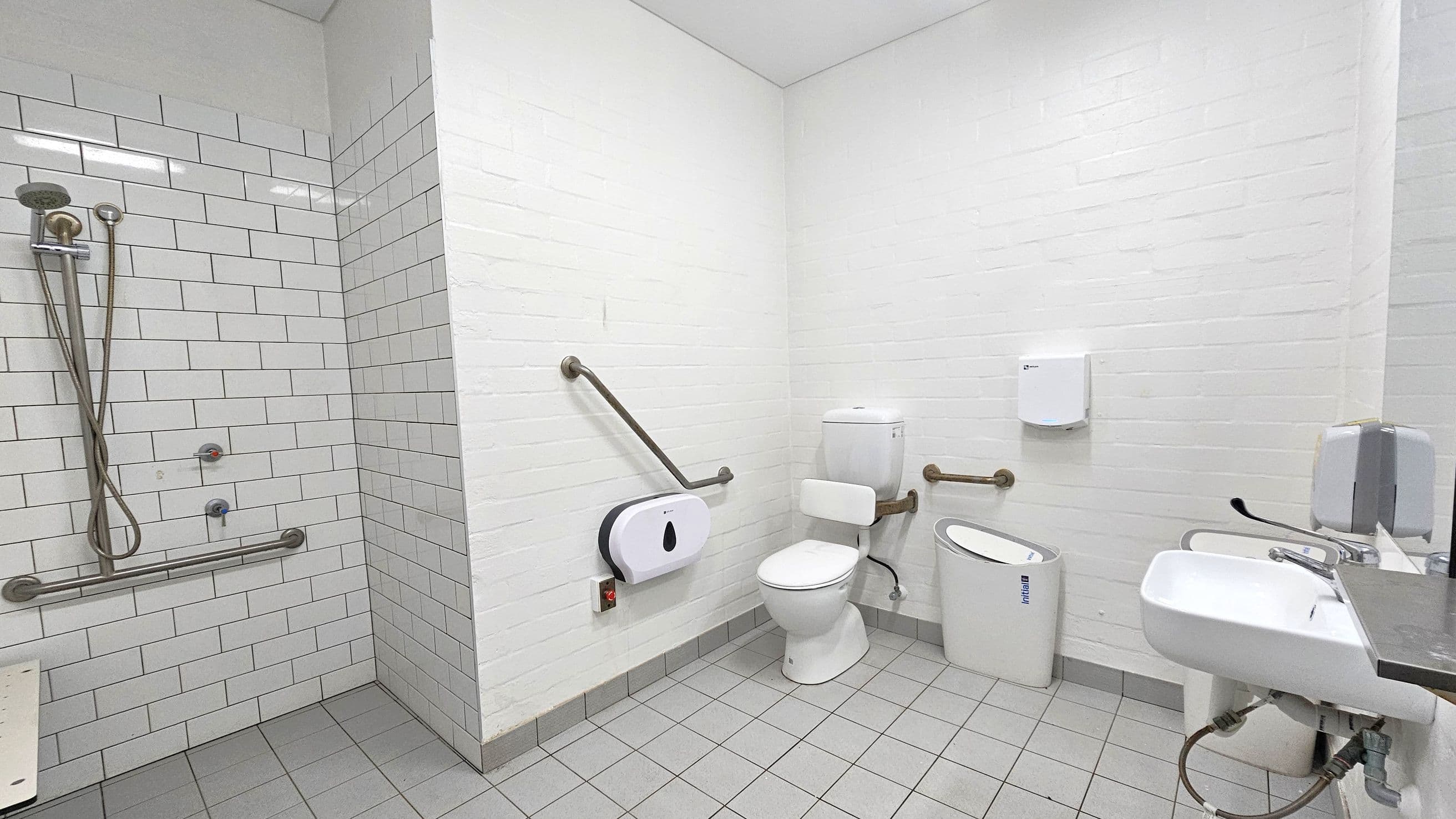 A white-tiled accessible bathroom with a shower area on the left, featuring a handheld showerhead and grab bars. In the centre, there's a toilet with support rails and a sanitary bin nearby. On the right, a small sink with a lever tap is mounted on the wall, accompanied by a soap dispenser. The floor is covered with grey tiles, and the ceiling is plain white.