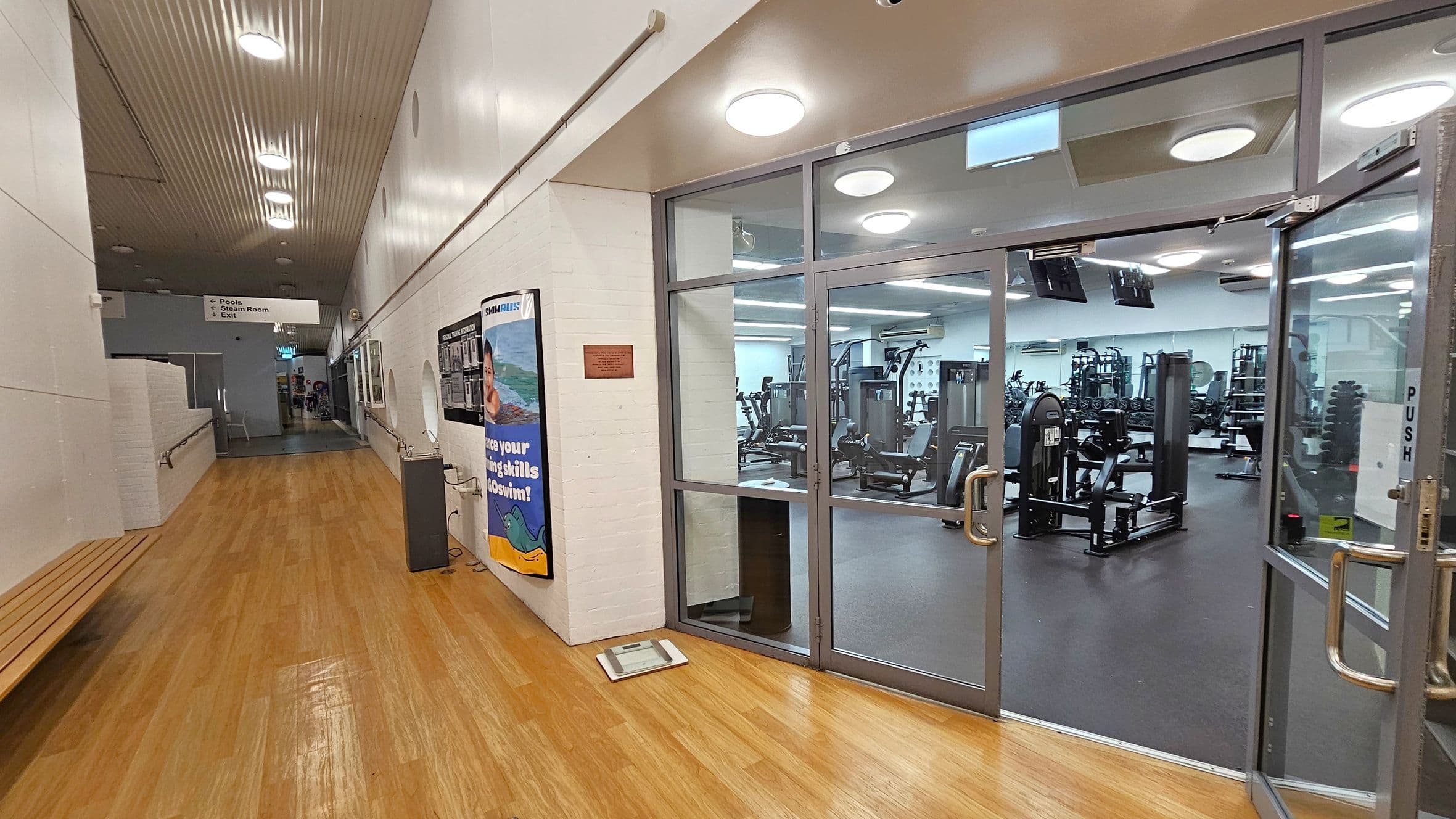 A hallway with a wooden floor and a white ceiling with recessed lights. On the right, there is a glass door leading to a gym with various exercise equipment visible inside. A poster is displayed on the wall next to the door. On the left, there is a wooden bench along the wall. Further down the hallway, a sign indicates directions to pools, a steam room, and an exit.
