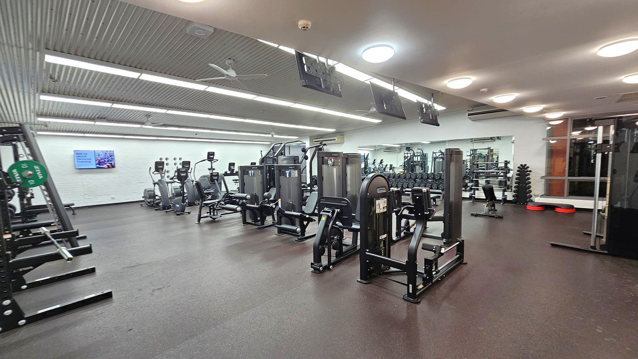 A spacious gym with various exercise machines. On the left, there are weightlifting racks with green weight plates. In the centre, several elliptical machines and strength training equipment are lined up. The right side features a mirrored wall reflecting more gym equipment, including dumbbells and benches. The ceiling has bright lights and a fan, while the floor is covered with dark rubber matting. A wall-mounted screen is visible in the background.