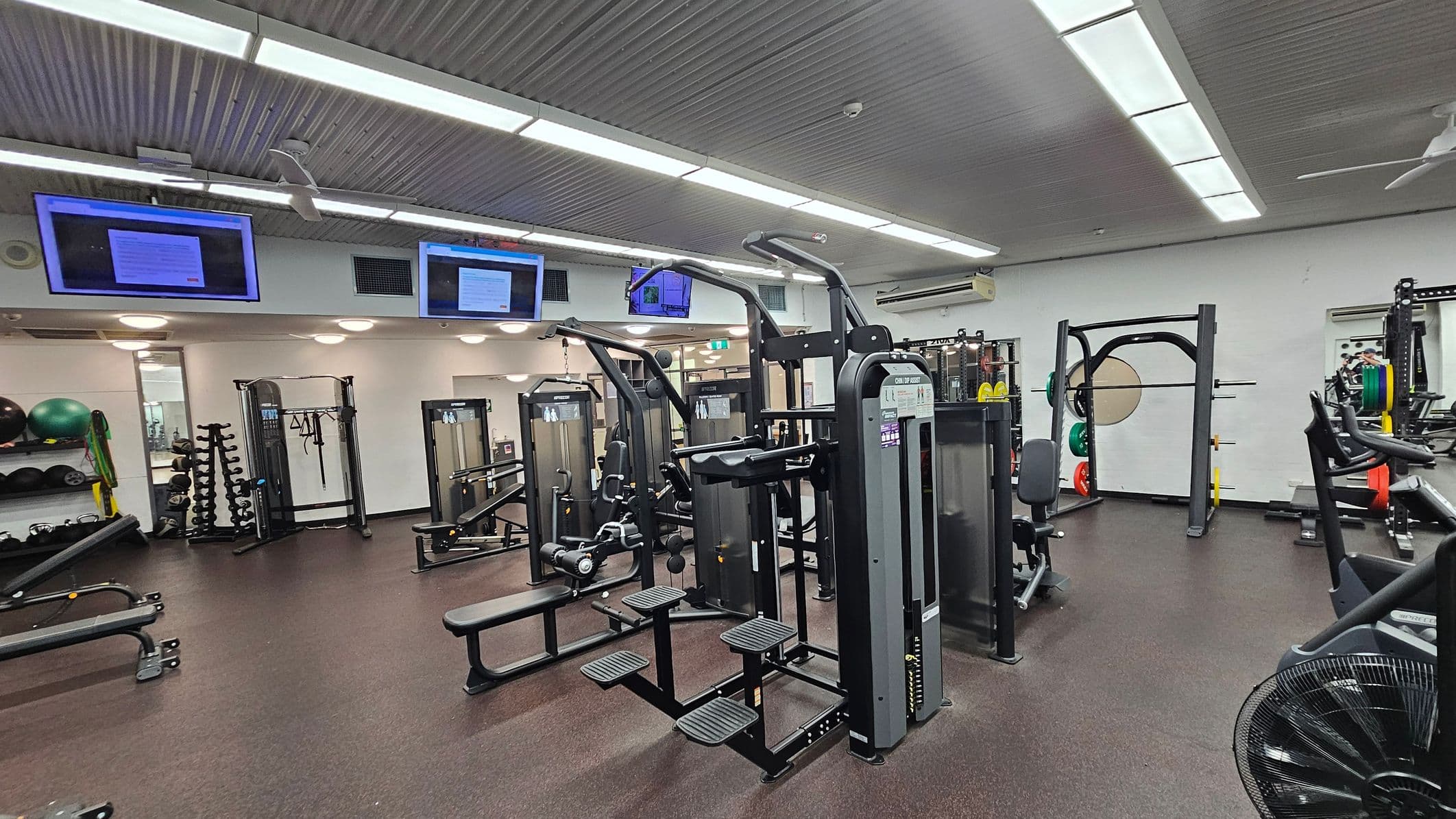 A gym with various exercise equipment. In the centre, there are weight machines and benches. On the left, shelves hold exercise balls and resistance bands, with a rack of dumbbells nearby. On the right, a squat rack with weight plates is visible. The ceiling has fluorescent lights and mounted screens displaying information. The floor is covered with dark rubber matting.