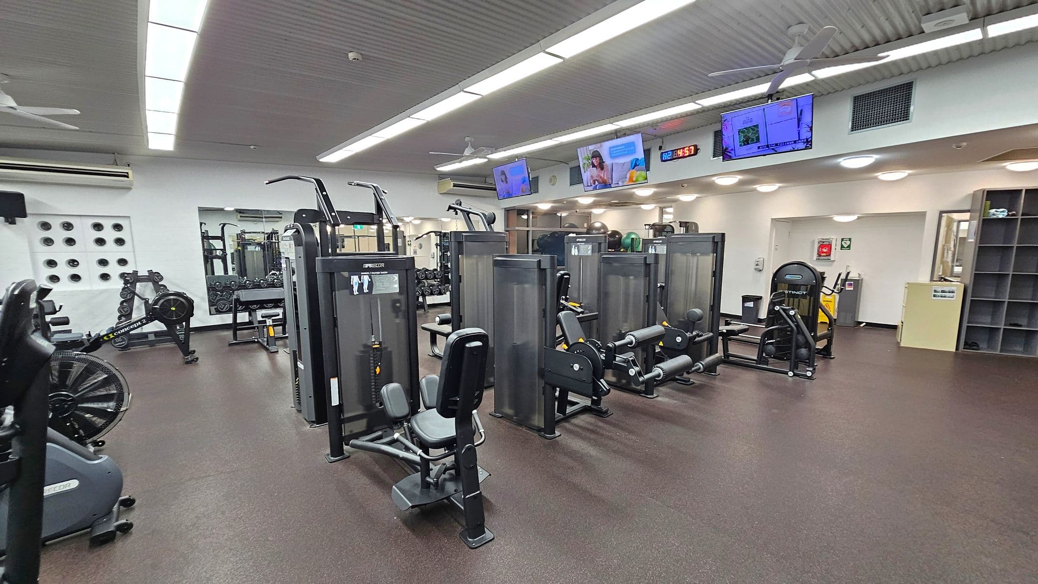 A gym with various exercise machines arranged in rows. On the left, there is a rowing machine and a wall with circular ventilation holes. In the centre, several weight machines are positioned, including leg and arm equipment. On the right, there are more machines and a shelving unit. The floor is dark with a speckled pattern, and the ceiling has fluorescent lights and ceiling fans. Several screens are mounted on the wall displaying different content.