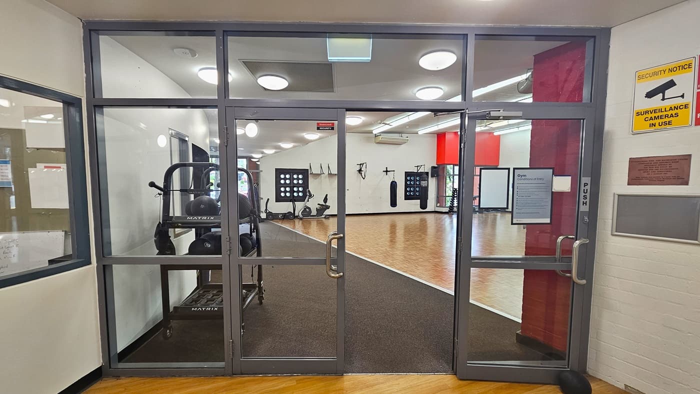 Glass doors with metal frames lead into a gym area. Inside, exercise equipment like ellipticals and punching bags are visible. The floor is wooden, and the ceiling has circular lights. On the right wall, there's a yellow sign indicating surveillance cameras are in use, along with a plaque. The left side shows a partial view of another room through a window.