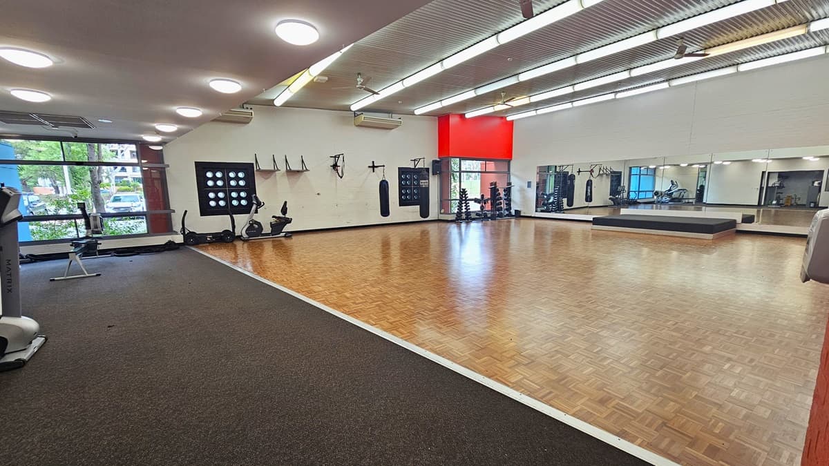 A spacious gym with a wooden floor in the centre and a carpeted area on the left. The left side features exercise equipment, including stationary bikes and a treadmill, near large windows. The right side has a mirrored wall and a raised platform. The ceiling is lined with fluorescent lights, and the walls are adorned with various fitness accessories. A red accent wall is visible in the background.