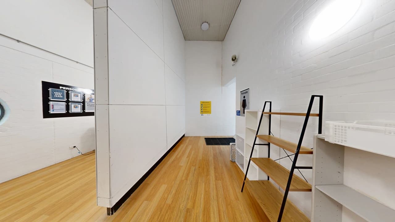 A room with light wooden flooring and white walls. On the right, there are two sets of shelves, one with a black frame and wooden shelves, the other white. In the centre, a black mat is on the floor near a door with a sign above it. On the left wall, a black-framed bulletin board displays various notices. The ceiling is light green with a circular light fixture.