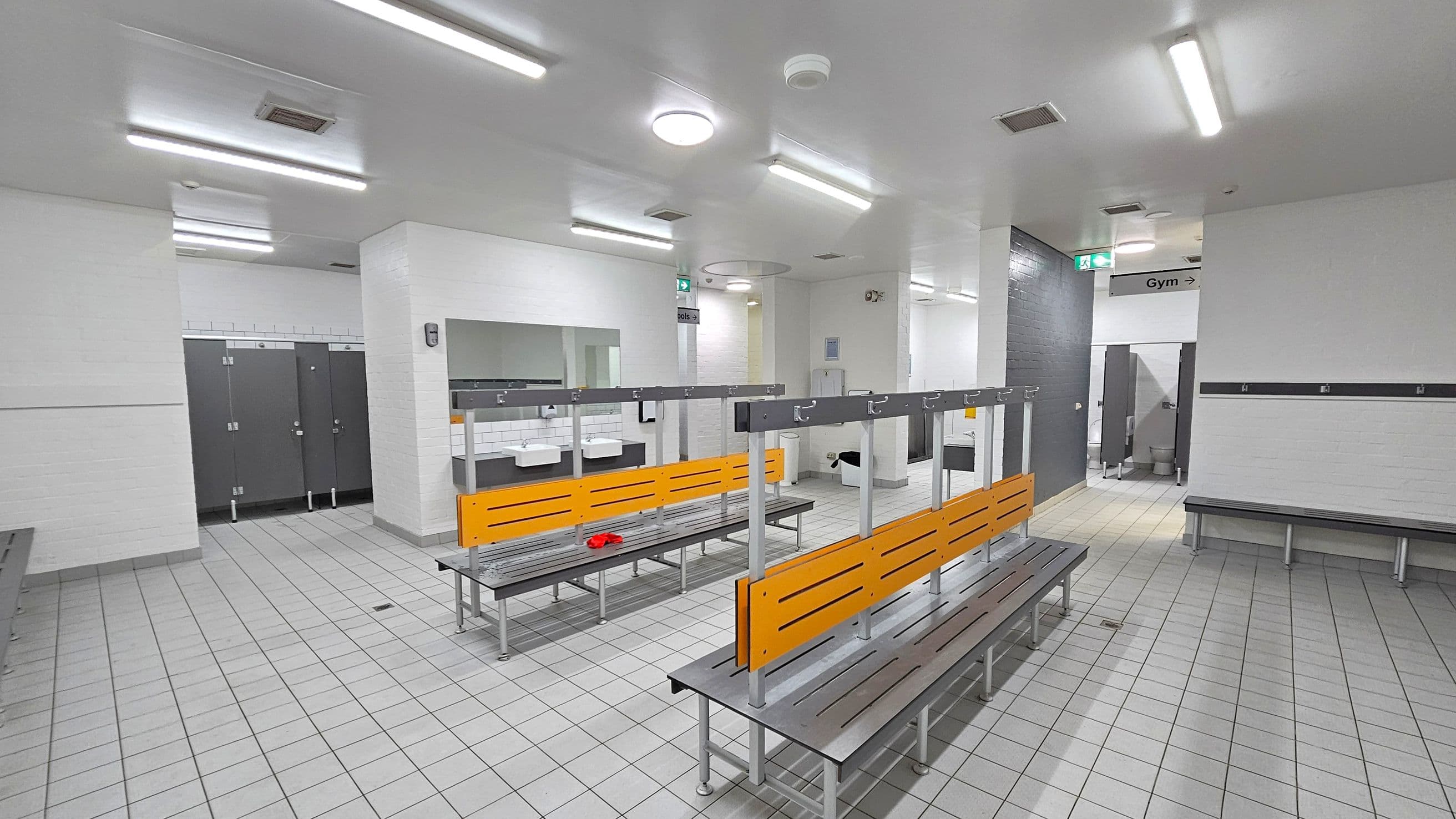 A spacious locker room with grey tiled flooring and white walls. In the centre, there are benches with orange backrests and metal hooks above. To the left, there are several closed grey lockers. In the background, sinks with mirrors are visible. On the right, a sign indicates the direction to the gym. The ceiling is white with fluorescent lights. A red item is placed on one of the benches.