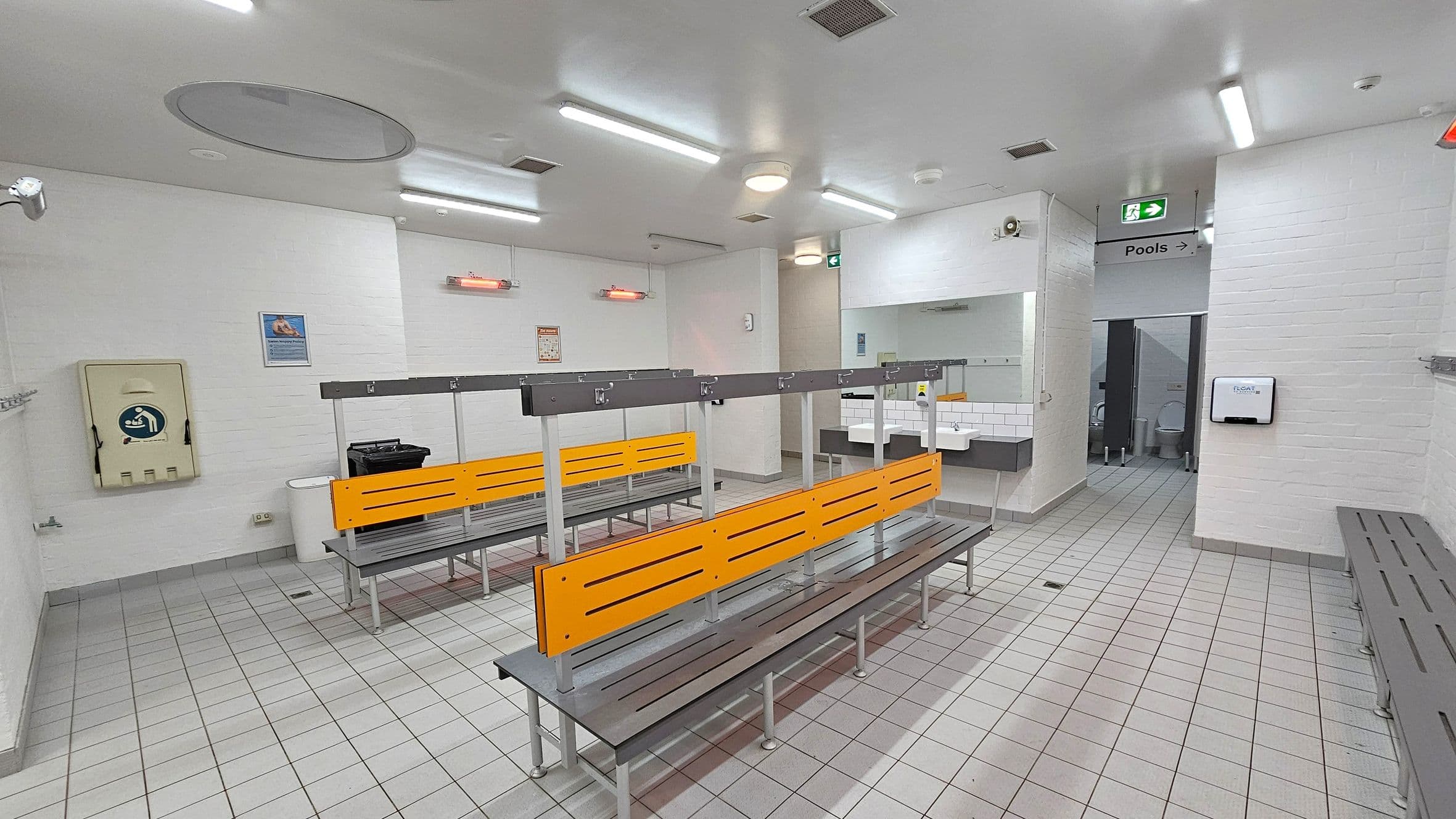A changing room with grey tiled flooring and a white ceiling with fluorescent lights. In the centre, there are benches with orange backrests and metal frames. On the left wall, there is a baby changing station and a waste bin. The right side has a sign indicating the direction to the pools and a glimpse of a toilet cubicle. Sinks with mirrors are visible near the entrance to the toilets. The walls are white brick, and there are coat hooks along the left wall.