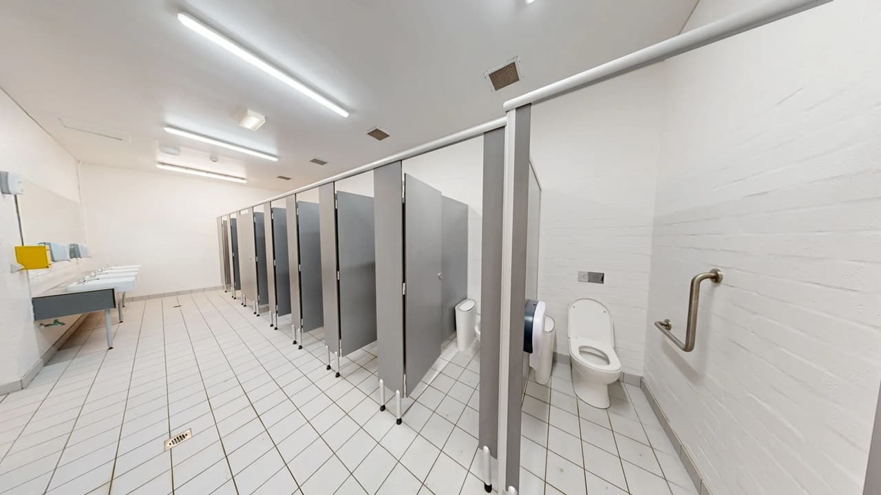 A public restroom with a row of toilet cubicles on the right, each with grey doors and partitions. The first cubicle door is open, revealing a white toilet and a grab bar on the wall. On the left, there are sinks with a yellow soap dispenser and a hand dryer. The floor is covered with white tiles arranged diagonally, and the ceiling has fluorescent lights. The walls are plain white, contributing to a clean and minimalistic appearance.