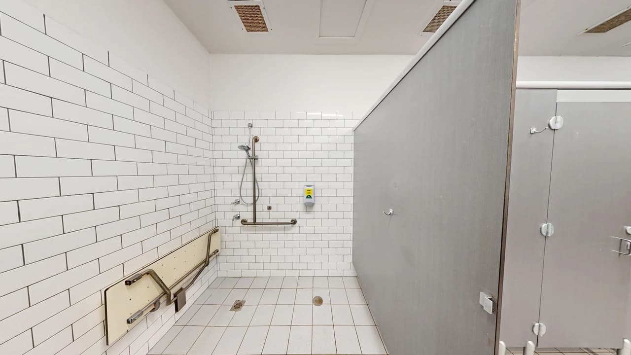 A bathroom with white subway tiles on the walls and floor. On the left, there is a foldable bench with metal support bars. In the centre, a showerhead is mounted on the wall with a grab bar below it. A soap dispenser is also visible. On the right, a grey partition separates the shower area from another section. The ceiling is plain white with ventilation panels.