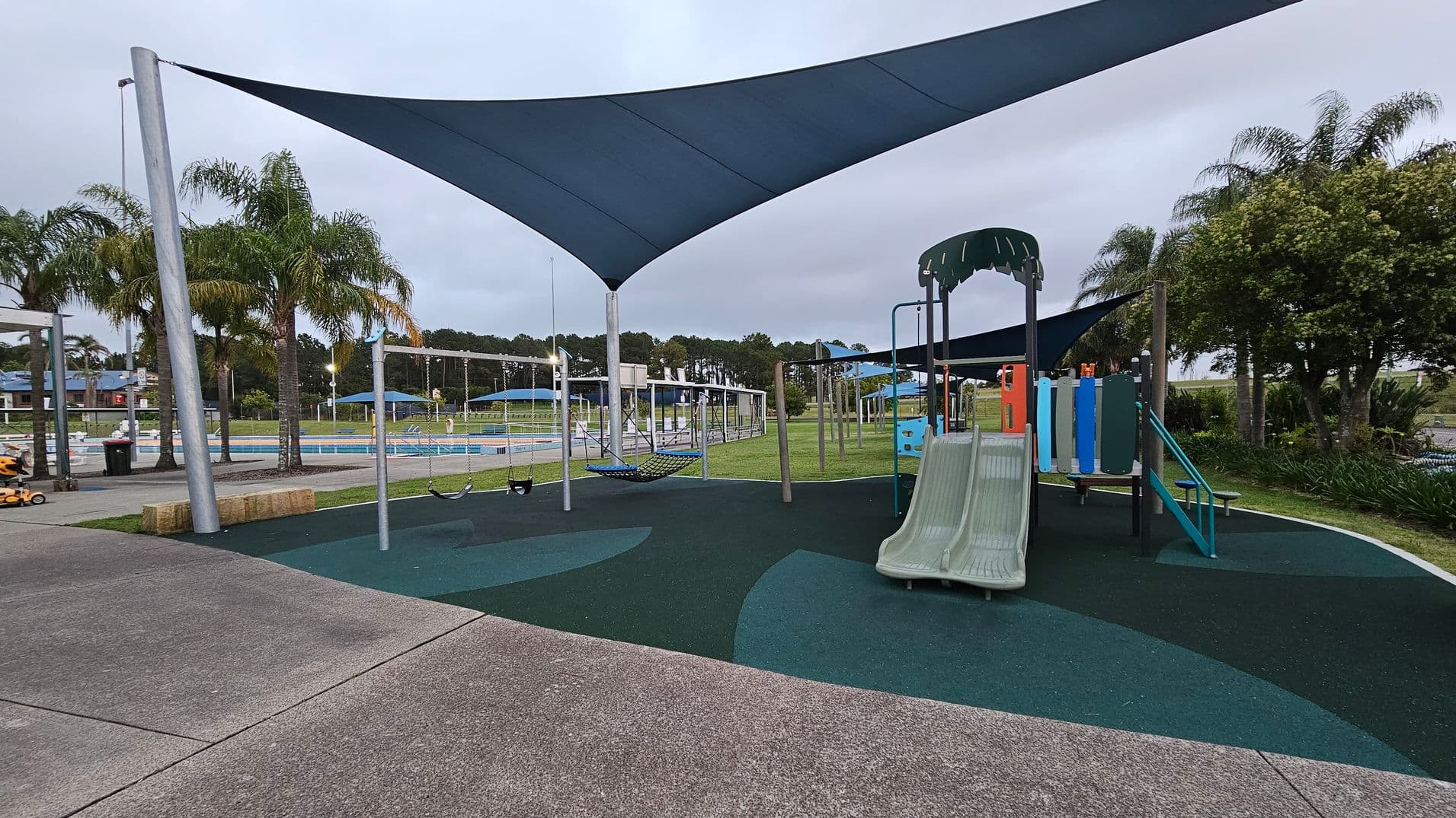 A playground with a slide and climbing structure on the right, featuring a green and blue colour scheme. In the centre, there are swings and a hammock under a large triangular shade sail. The ground is covered with a rubber safety surface in green and blue patterns. On the left, there is a concrete path and some palm trees. In the background, a fenced area with a pool and more shade structures is visible, along with more trees and grass.