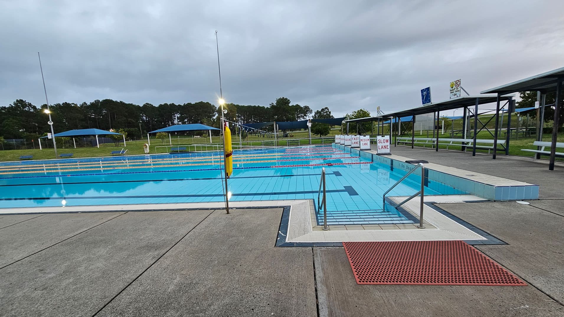 Outdoor swimming pool with clear blue water, divided into lanes marked as "Fast Lane," "Medium Lane," and "Slow Lane." A metal handrail leads into the pool. Surrounding the pool is a concrete deck with a red mat near the entrance. In the background, there are green lawns, trees, and blue shade structures. Benches and a covered seating area are visible on the right. The sky is overcast, and pool lights are on.