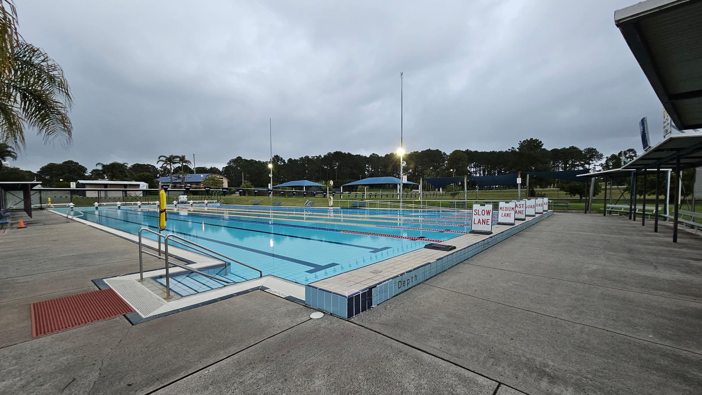 An outdoor swimming pool with multiple lanes, each marked with signs for different speeds: "Slow Lane," "Medium Lane," and "Fast Lane." The pool is surrounded by a concrete deck. On the left, there are palm trees and a grassy area. In the background, there are covered seating areas and trees. The pool has metal handrails and a red mat at the entrance. Overhead lights illuminate the area.