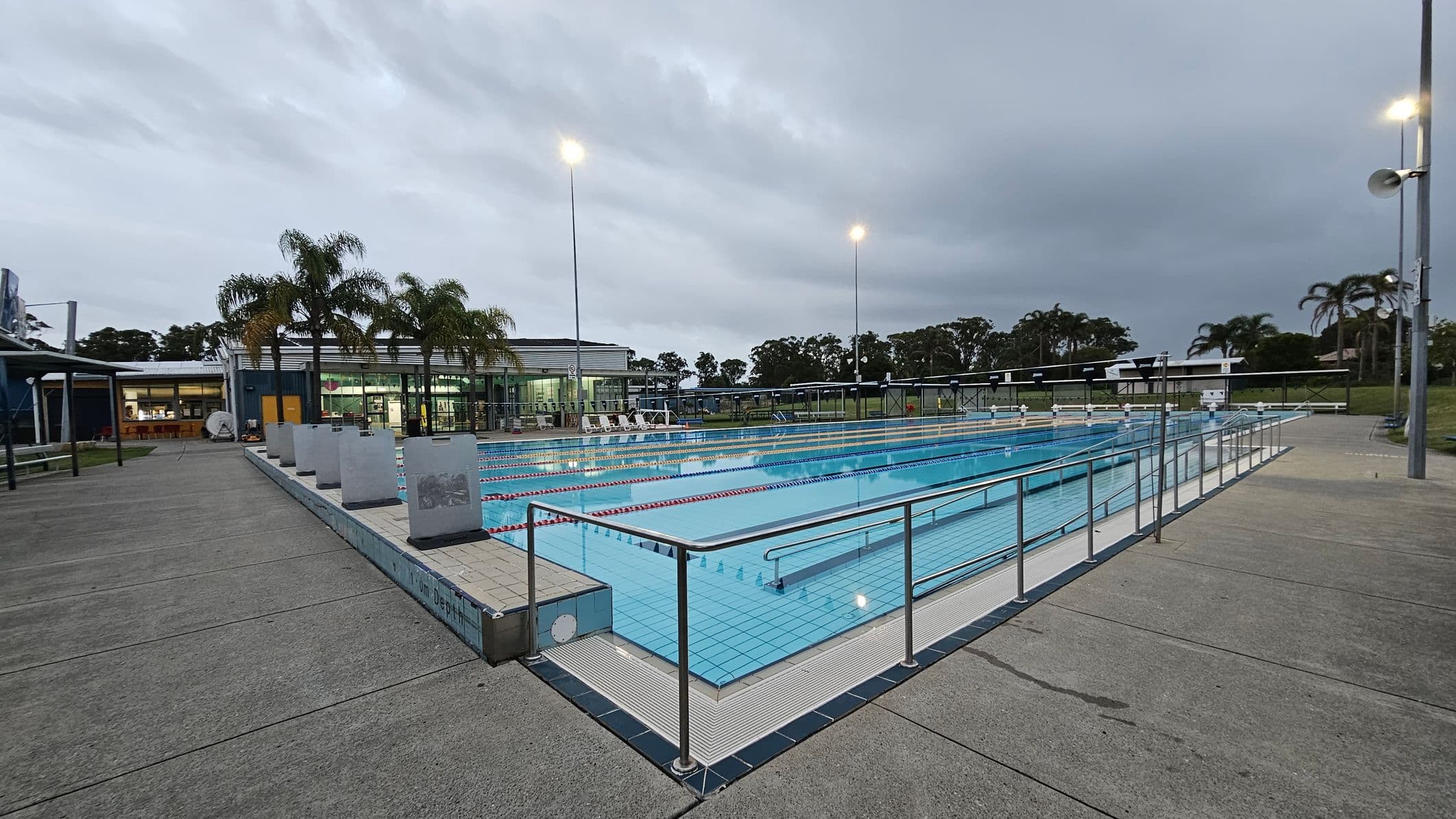 An outdoor swimming pool with multiple lanes, surrounded by a metal railing. The pool area is bordered by a concrete deck. On the left, there are palm trees and a building with large windows, possibly a facility or changing rooms. In the background, more palm trees and a grassy area are visible. Tall light poles illuminate the scene.