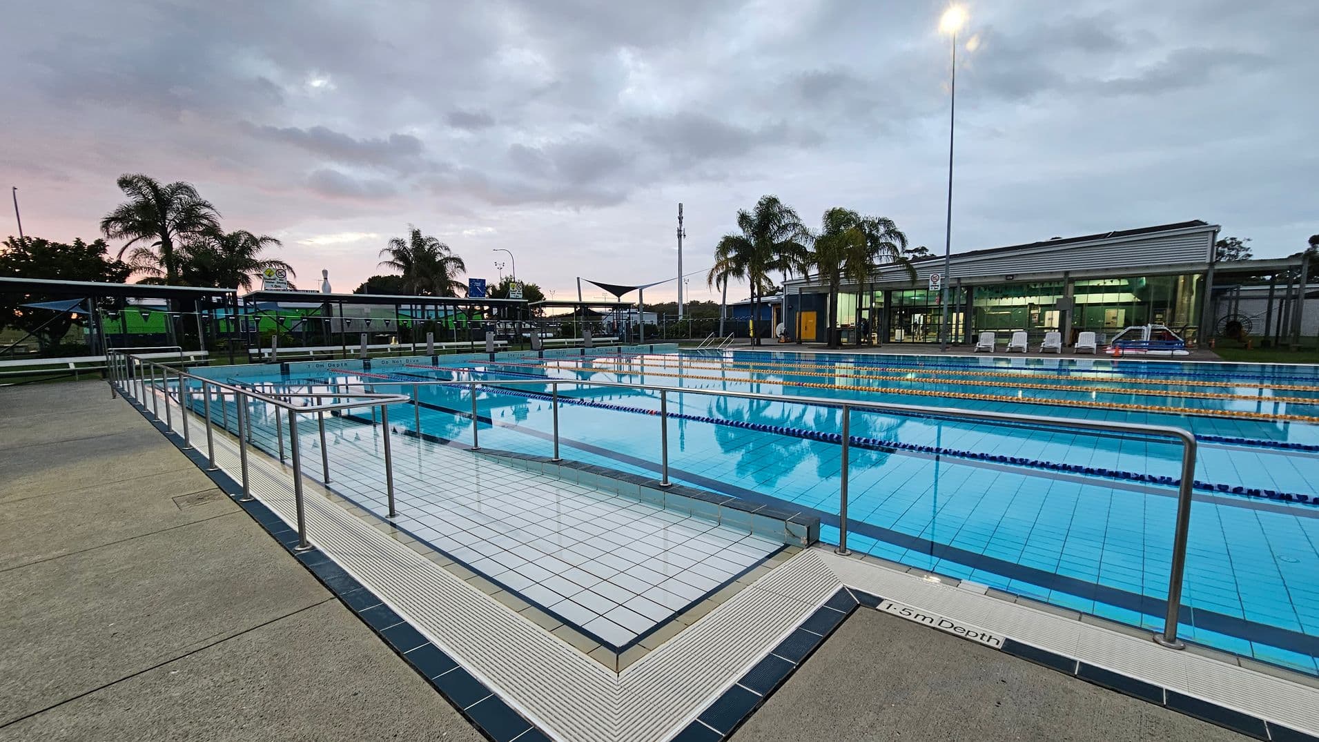 Outdoor swimming pool with clear blue water, surrounded by metal railings. The pool has lane dividers and a marked depth of 1.5 metres. On the right, a modern building with large windows and a few white lounge chairs is visible. Palm trees are scattered around the area, and a tall light pole is illuminated. The ground is paved with light-coloured tiles.