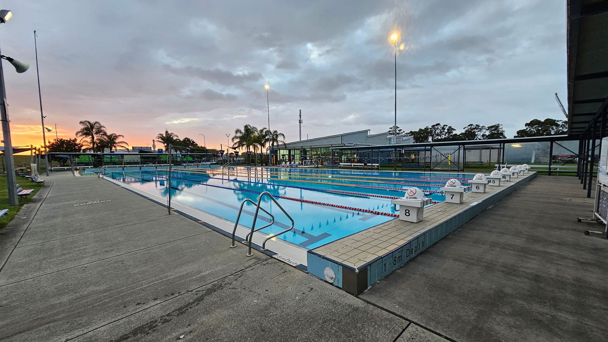 An outdoor swimming pool with eight lanes, each marked by a starting block. The pool is surrounded by a concrete deck. On the left, there are palm trees and a grassy area with benches. In the background, a building with large windows is visible. The sky is overcast with a hint of sunset on the horizon. Tall floodlights illuminate the area.