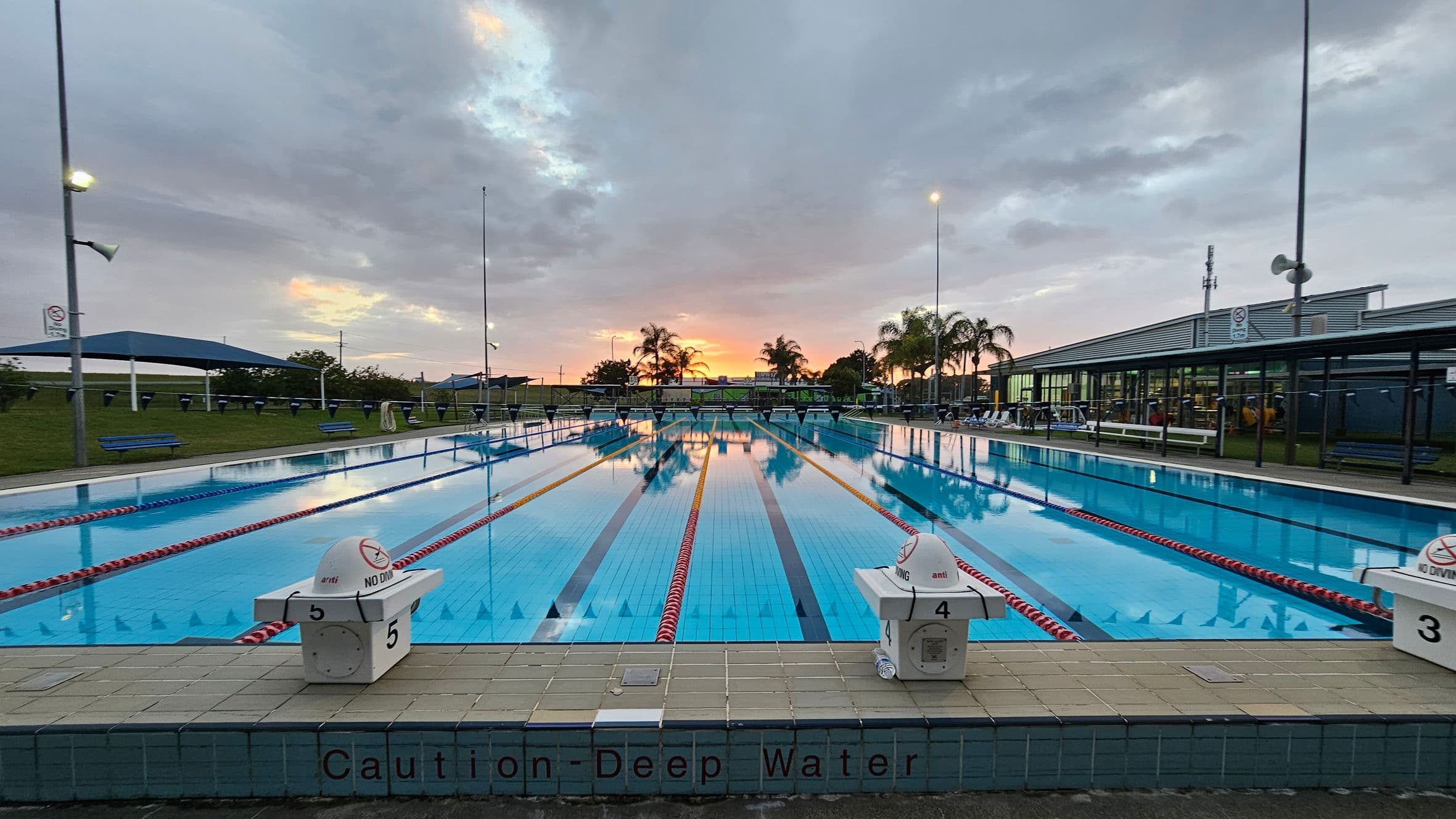 An outdoor swimming pool with marked lanes stretches into the distance. On the left and right, there are diving blocks labelled with numbers and "No Diving" signs. The pool is surrounded by a tiled deck with a "Caution - Deep Water" warning. In the background, palm trees and a building with large windows are visible. The sky is overcast with a hint of sunset colours. Benches and shaded areas are on the left, and a grassy area is visible beyond the pool.