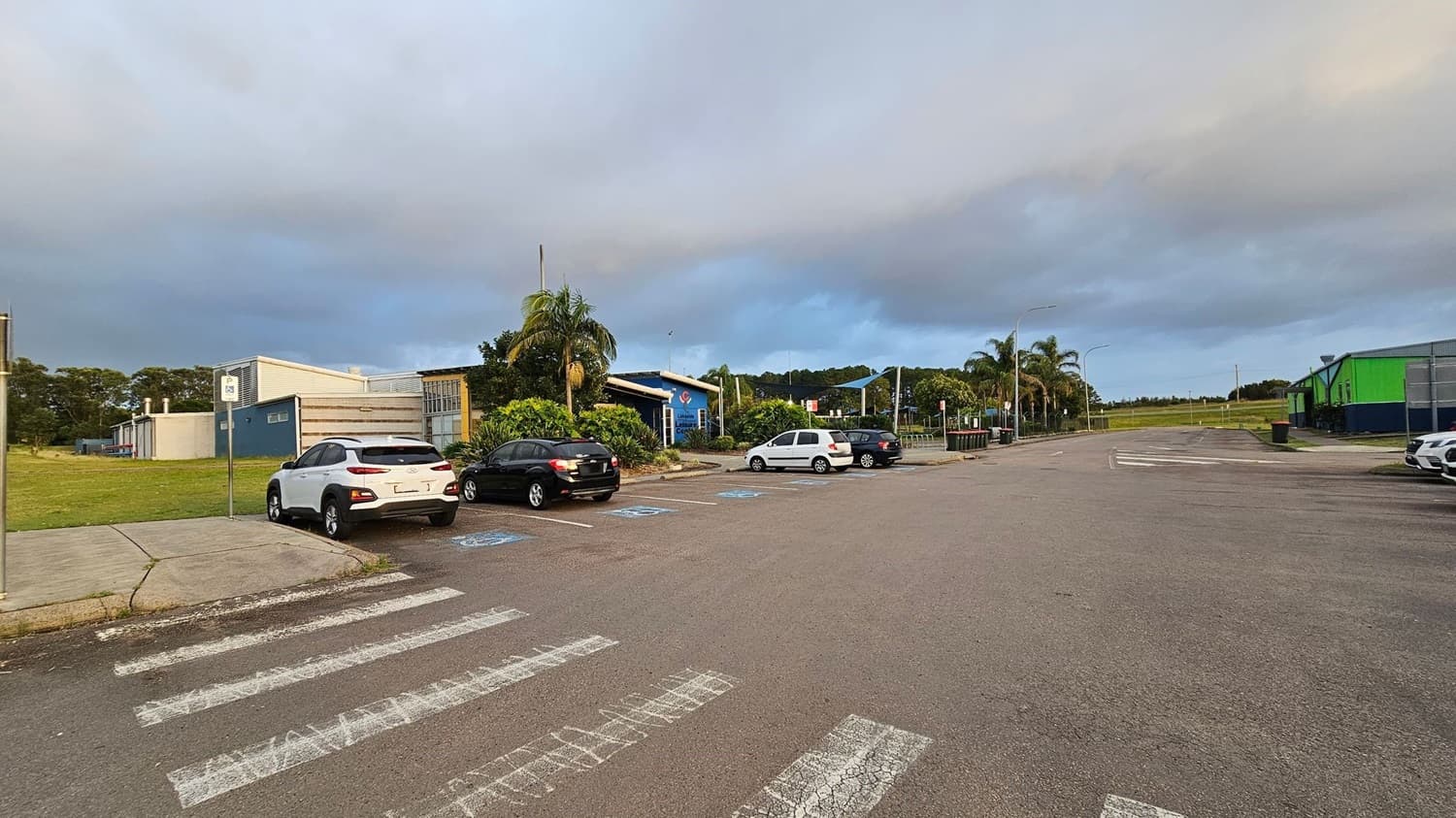 A paved road on the left with parked cars in the distance, lined with street lamps and trees, including palm trees. On the right, a grassy area is enclosed by a chain-link fence, featuring a structure with a blue roof and white poles. The background includes more trees and a few buildings.