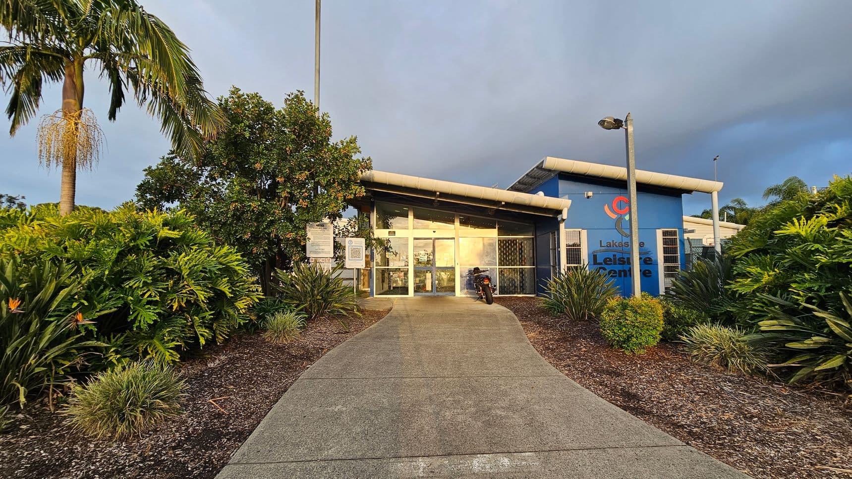 A building with a glass entrance is in the centre, with a path leading up to it. On the right, there's a blue wall with the words "Lakeside Leisure Centre" and a logo. A motorcycle is parked near the entrance. The foreground has a variety of green plants and shrubs, with a palm tree on the left. Two tall lamp posts are positioned near the entrance. The building has a slanted roof, and the area is surrounded by lush greenery.