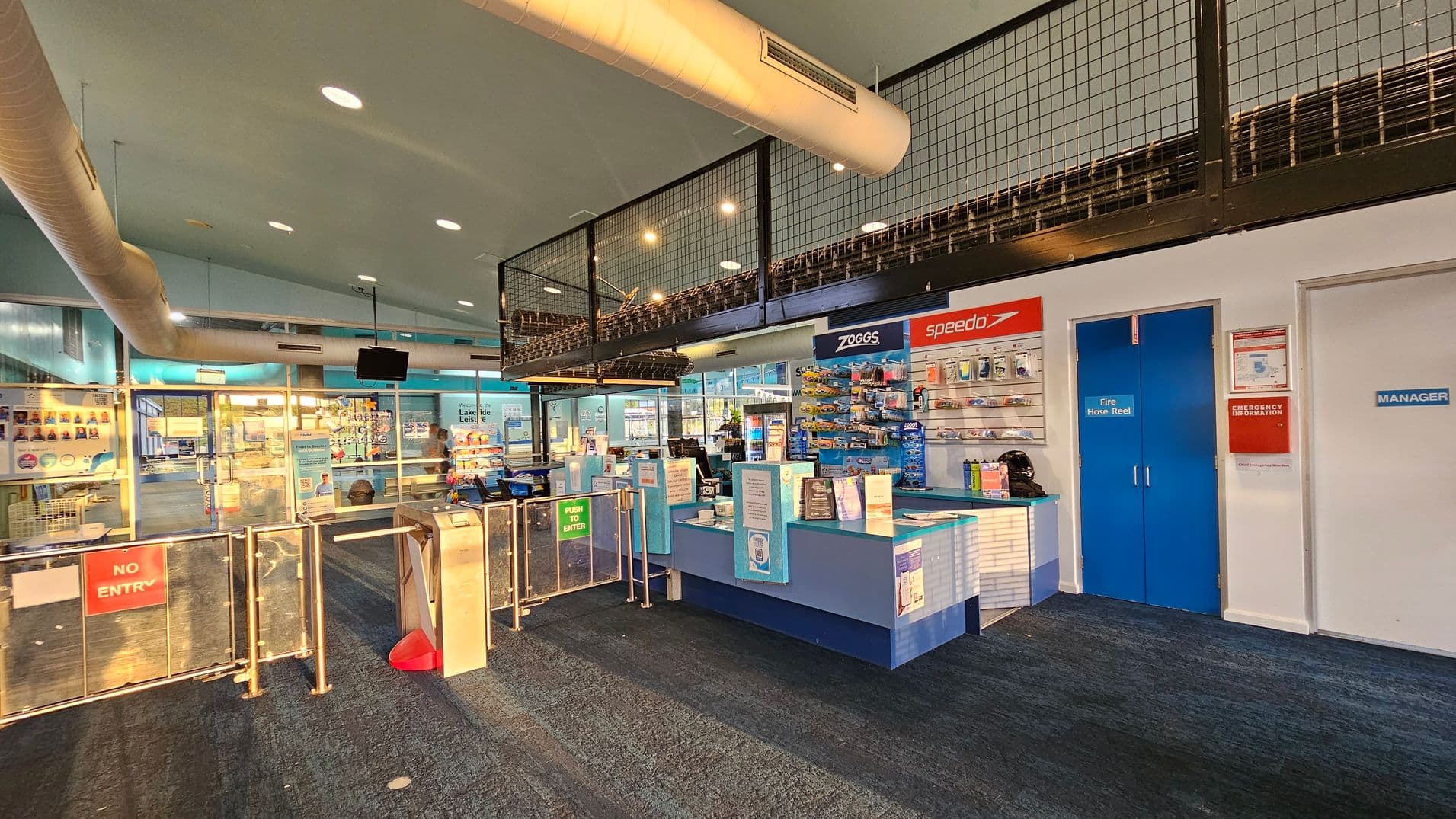 Entrance to a building with glass doors. On the left, the door displays the Port Stephens logo and several stickers. On the right, a large blue sign lists conditions of entry and facility opening hours. Inside, there are turnstiles and a reception area. A motorcycle is partially visible in the lower right corner. The floor is dark and the ceiling has lights and air conditioning vents.