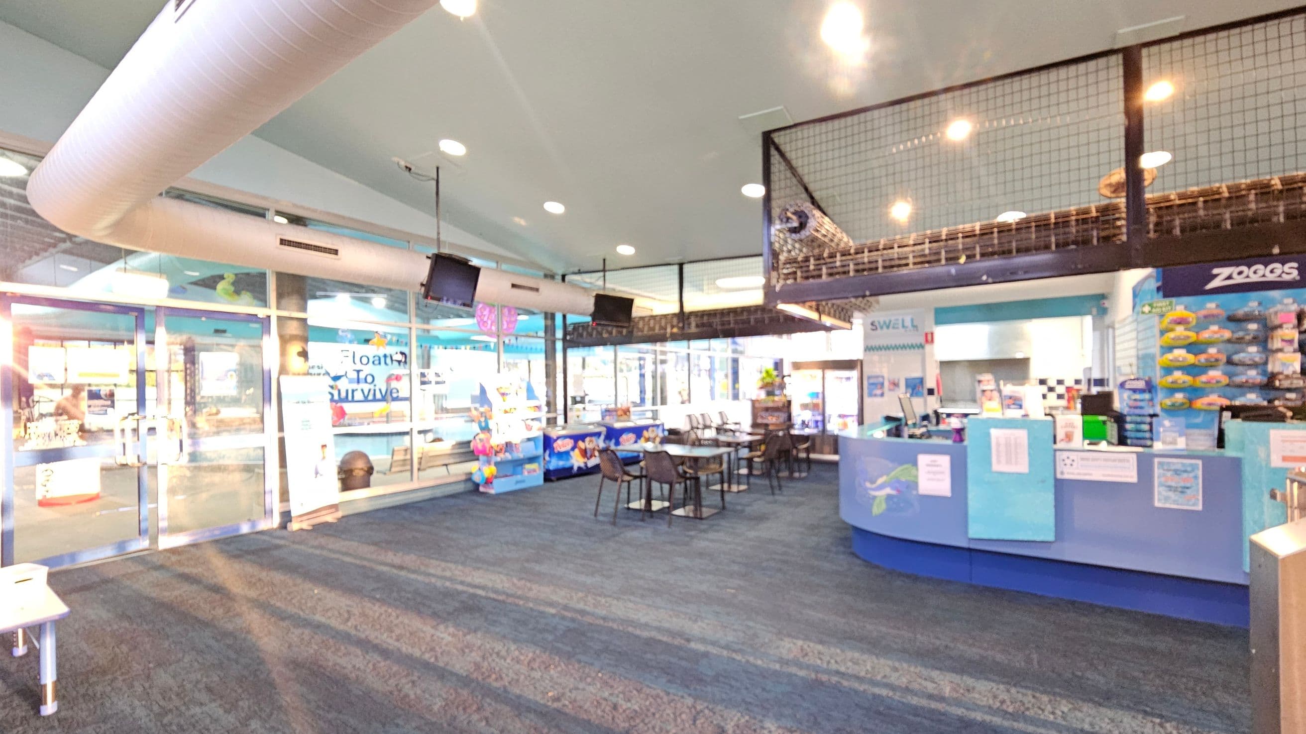 A spacious indoor area with a blue-themed reception desk on the right, displaying various swimming products. In the centre, there are tables and chairs for seating. The left side features large glass doors and windows with signage, allowing natural light to enter. Above, a netted mezzanine level is visible. The ceiling has recessed lighting and a large white air duct. The floor is carpeted in shades of blue and grey.