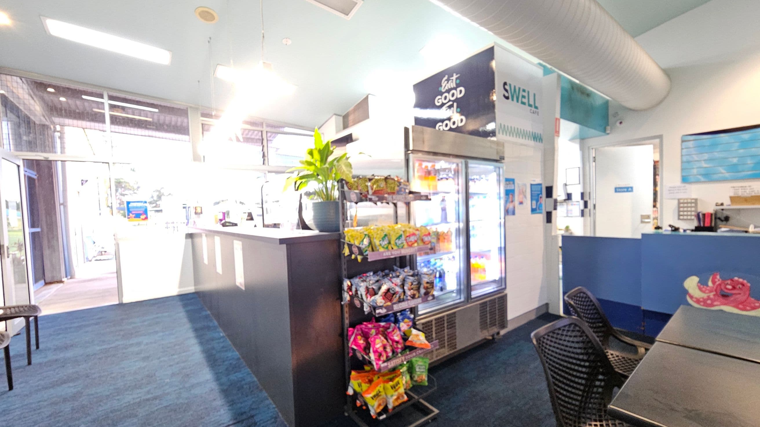 Inside a café with a blue carpeted floor. On the left, there's a glass entrance with natural light streaming in. In the centre, a counter with a plant on top and a snack display below. Behind it, a refrigerator stocked with drinks. On the right, a seating area with black chairs and tables. Above, a large duct runs along the ceiling. A sign reads "SWELL Café" and a colourful octopus design is visible on the counter.