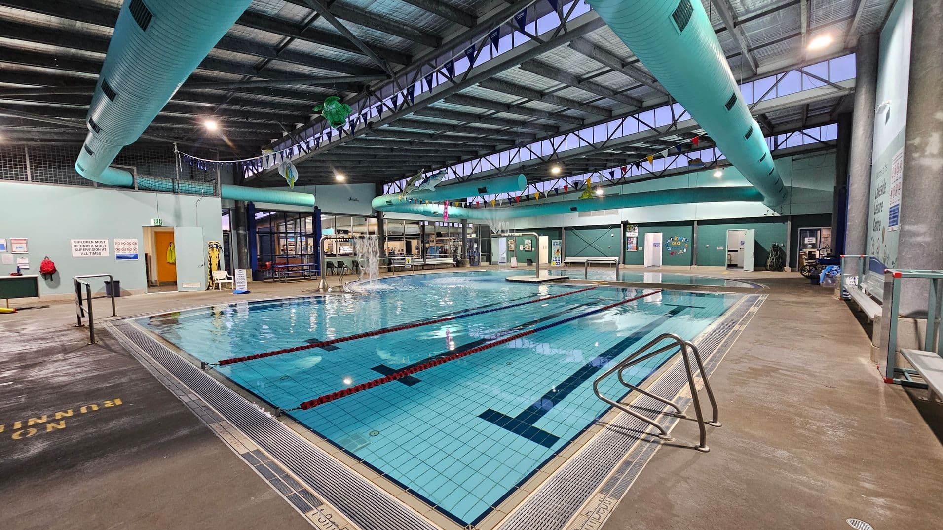 Indoor swimming pool with clear blue water, marked lanes, and a metal handrail on the right. The pool is surrounded by a concrete floor with safety signs. On the left, there are doors and a notice board. The ceiling features large turquoise ventilation ducts and exposed beams. In the background, there are windows, a seating area, and a small café or reception area. Bunting hangs across the space, adding a decorative touch.