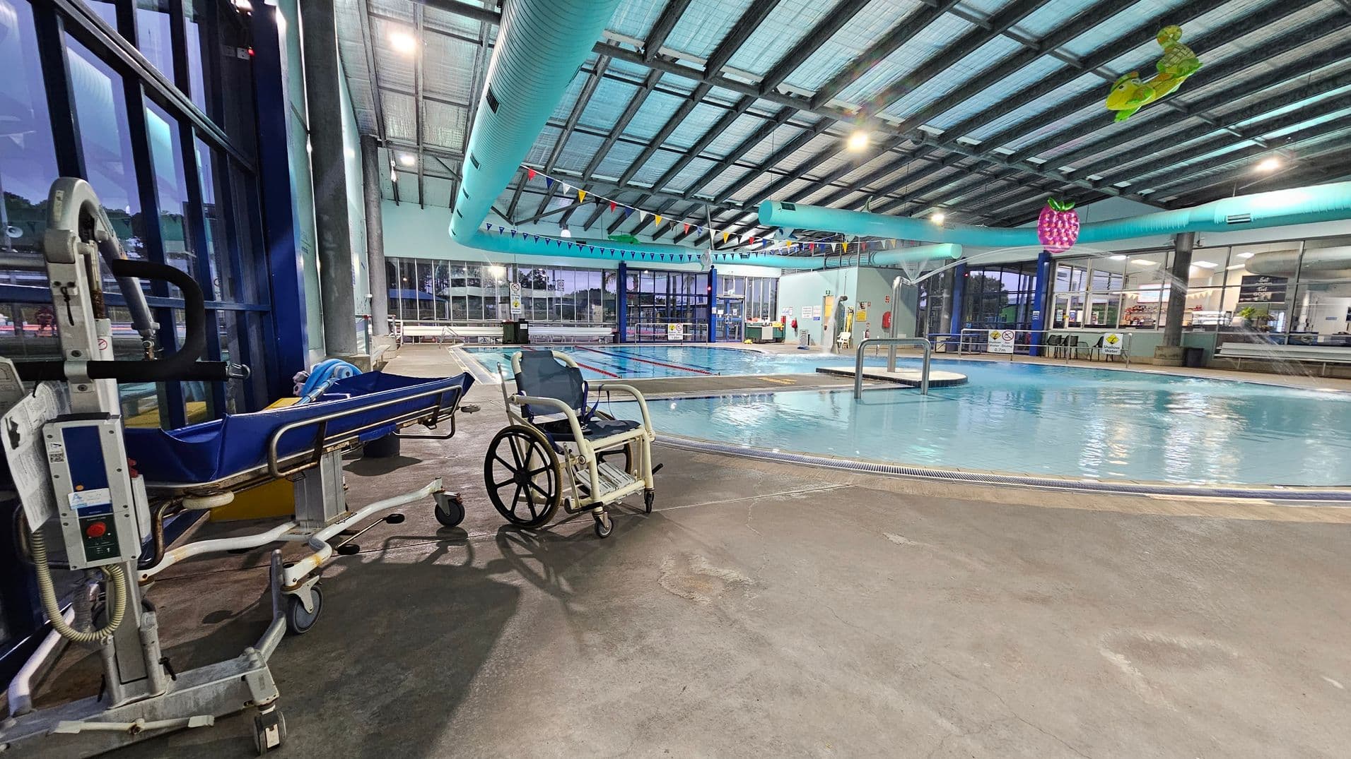 An indoor swimming pool area with a concrete floor. On the left, there is a blue medical stretcher and a wheelchair. The pool is in the centre, surrounded by a railing, with a ramp leading into the water. Colourful triangular flags hang above the pool. The ceiling is supported by metal beams and features large blue ventilation ducts. In the background, there are large windows and a seating area with tables and chairs.