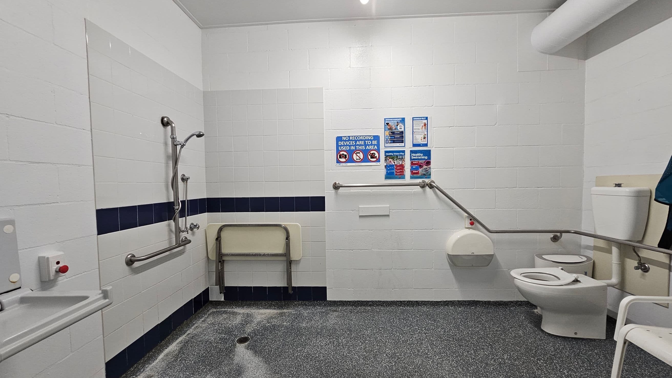 A public bathroom with white tiled walls and a speckled grey floor. On the left, there is a shower area with a metal grab bar and a foldable seat. In the centre, several signs are displayed on the wall, including a "No Recording Devices" notice. On the right, there is a toilet with a grab bar and a toilet paper dispenser. A white ceiling with a light fixture is visible.