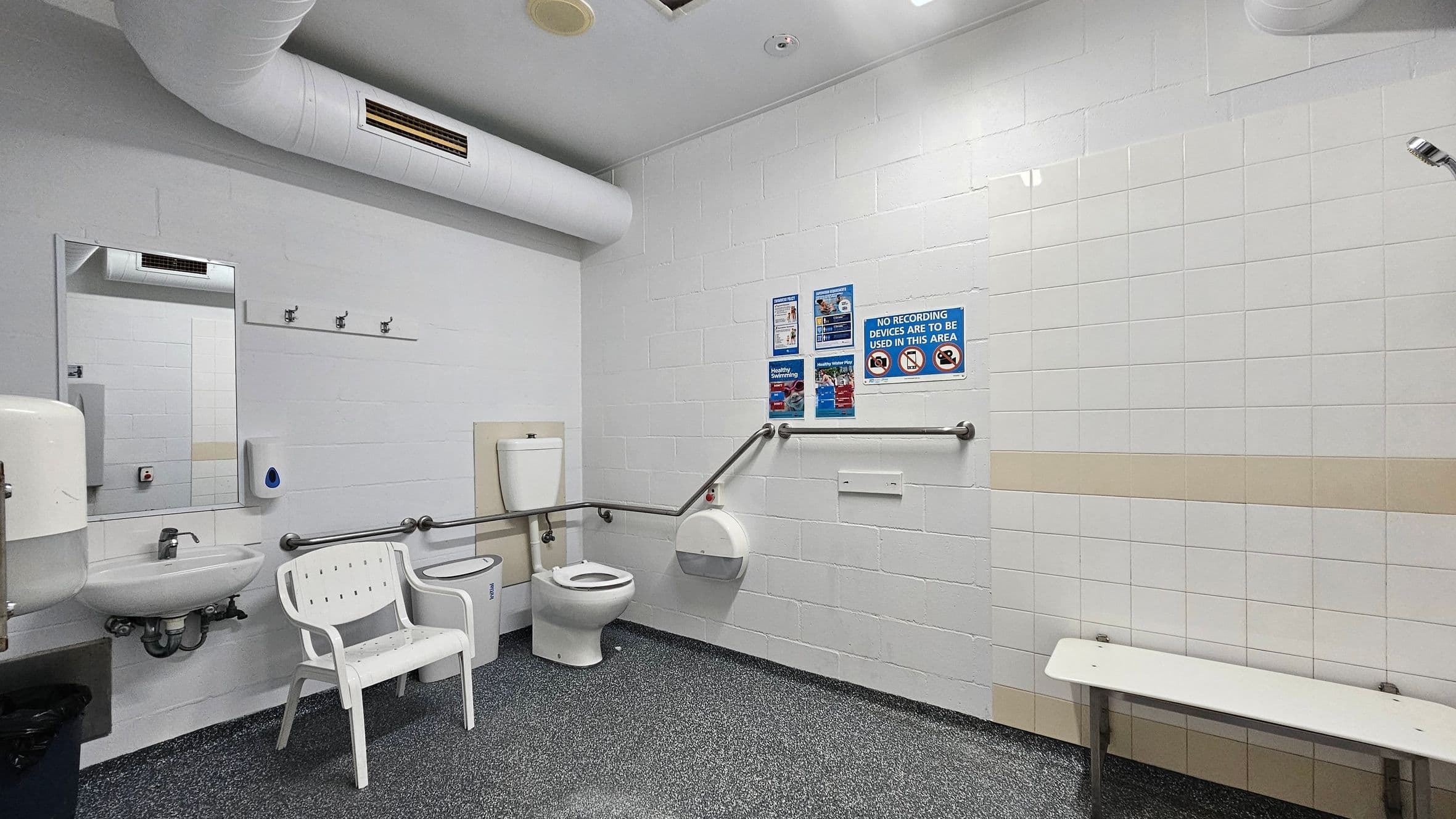 A spacious, accessible bathroom with white tiled walls and a speckled grey floor. On the left, there's a sink with a mirror above it, a soap dispenser, and a white chair. A toilet with grab rails is in the centre. On the right, a white bench is attached to the wall. Several informational signs are posted above the toilet. The ceiling has exposed ductwork and lighting fixtures.