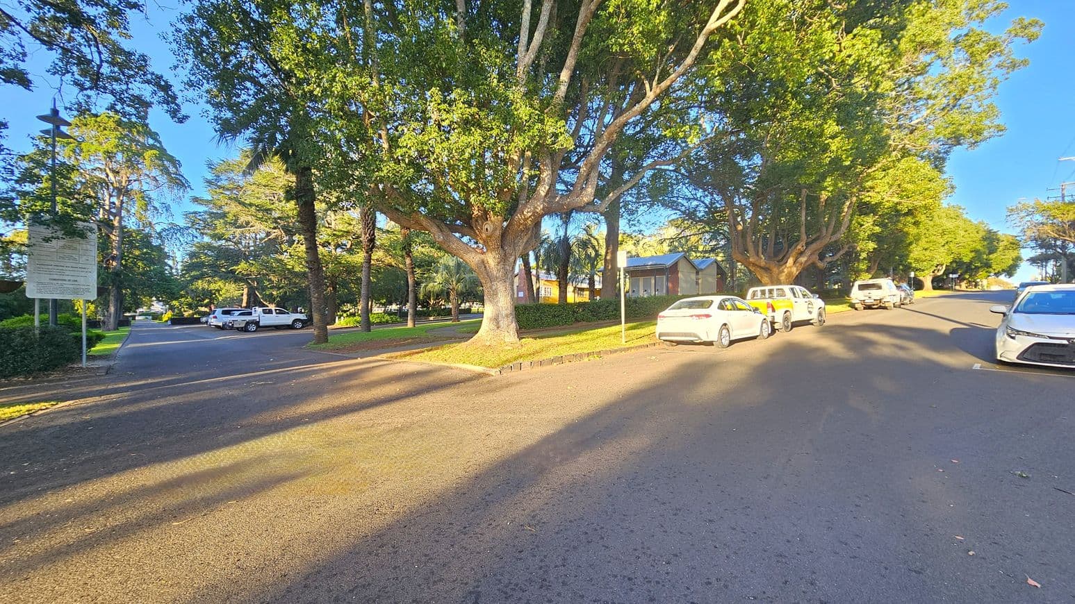 A street scene with large trees lining both sides. On the left, a signpost and a few parked cars are visible near a pathway leading into a park-like area. On the right, several cars are parked along the kerb. A small building with a sloped roof is partially visible among the trees. The road is wide and clear, with sunlight casting shadows from the trees. The area is lush with greenery, and the sky is clear.
