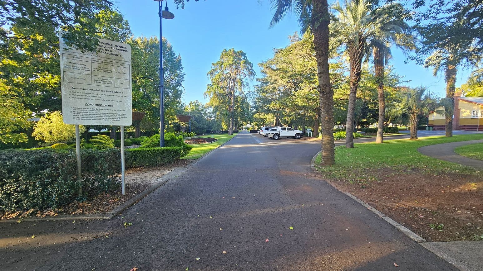 A paved road runs through a park with a large sign on the left detailing parking arrangements. To the right, several palm trees line a grassy area with a curved path. In the background, two white vehicles are parked on the right side of the road. The area is surrounded by lush green trees and well-maintained gardens. A streetlamp is visible on the left, and a building is partially visible on the right.