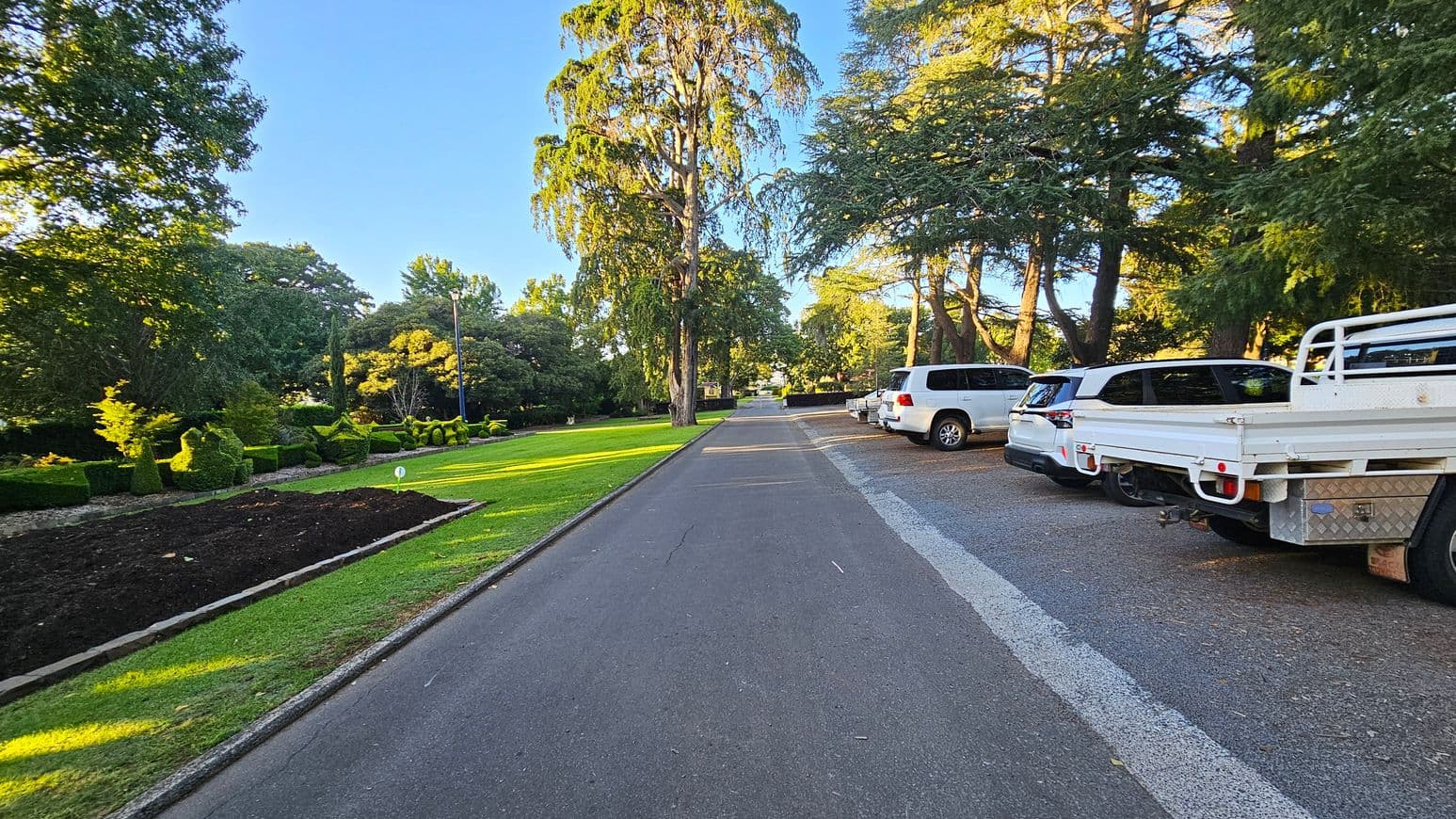 A paved road runs through the centre, with a row of parked vehicles, including a white truck, on the right. On the left, there is a well-maintained garden with neatly trimmed hedges and a small patch of dark soil. Tall trees line both sides of the road, casting shadows on the grass and pavement. The scene is well-lit, suggesting daylight, and the road stretches into the distance, flanked by greenery.
