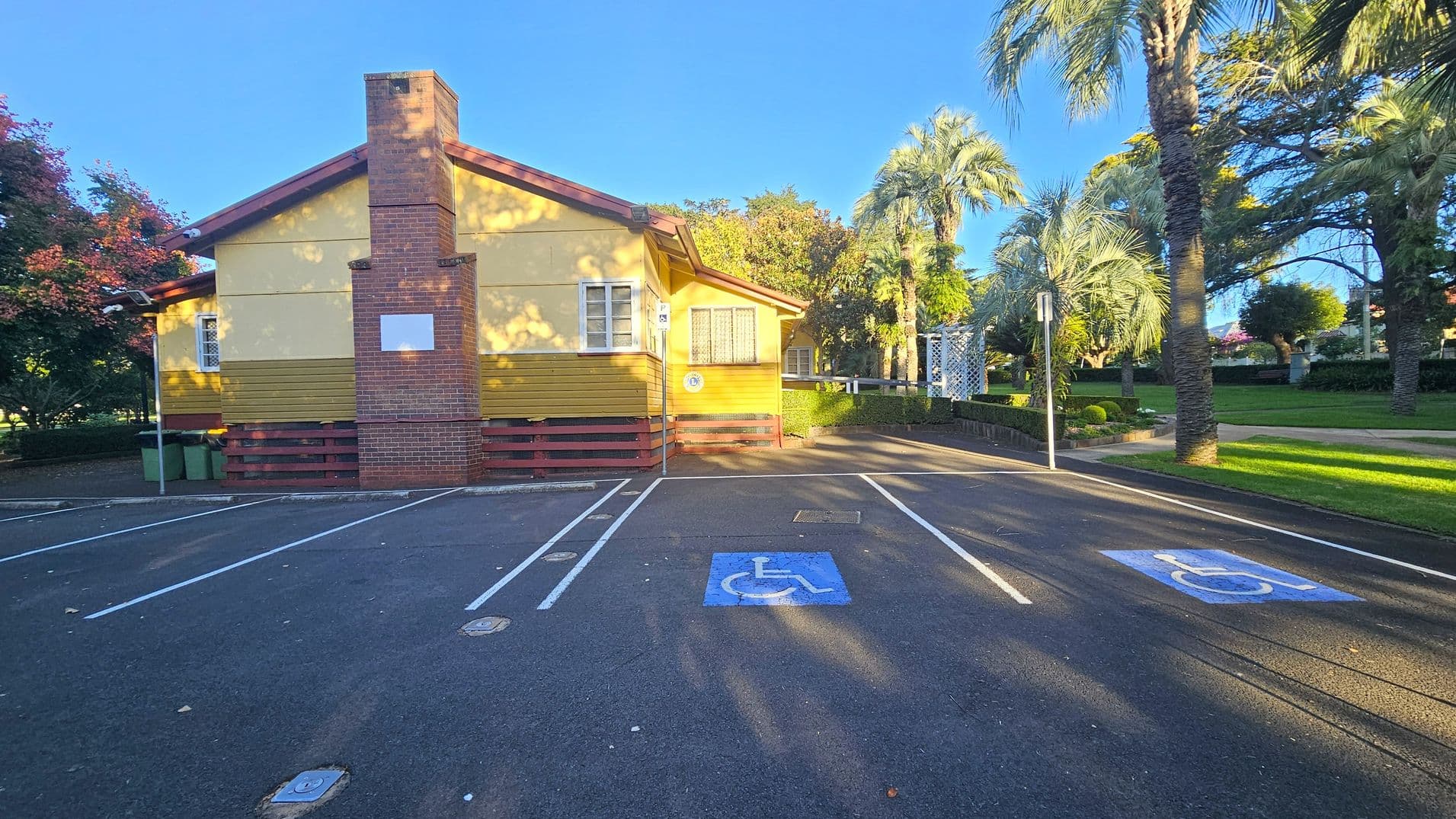 A yellow and brown building with a prominent brick chimney is in the centre. The foreground features a car park with marked disabled parking spaces. On the right, there are palm trees and a grassy area with a pathway. To the left, there are green bins and a tree with red leaves. The background includes more trees and a hedge. The scene is well-lit, suggesting daylight.